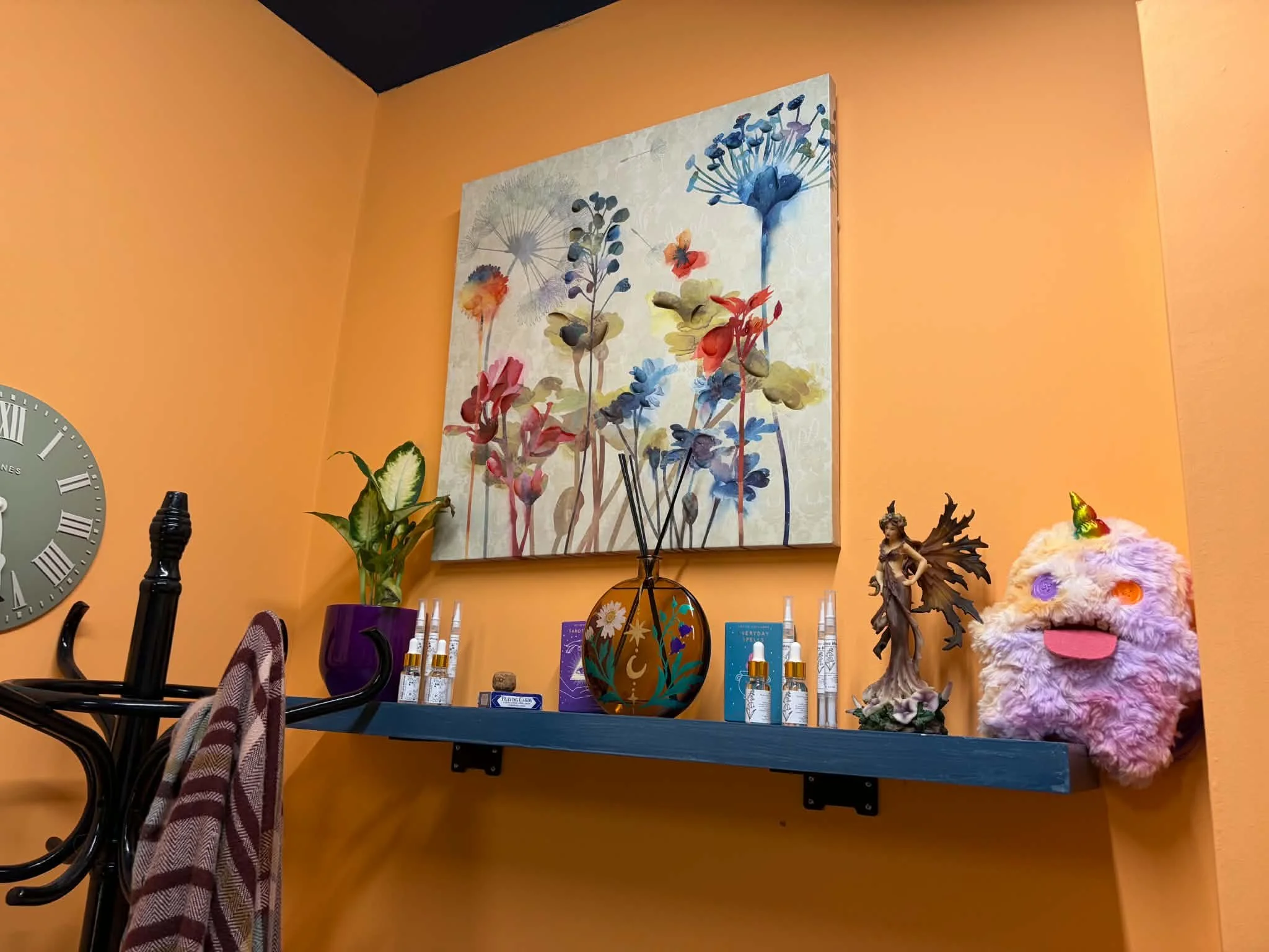 A decorated display area with a floral painting, small decorative figurines, a purple flower pot, various bottles, a clock, and a plush teddy bear on an orange wall.