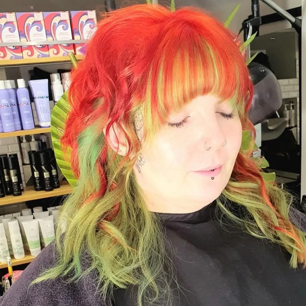 Woman with vibrant red, yellow, and green hair, sitting in a salon with shelves of hair products in the background.