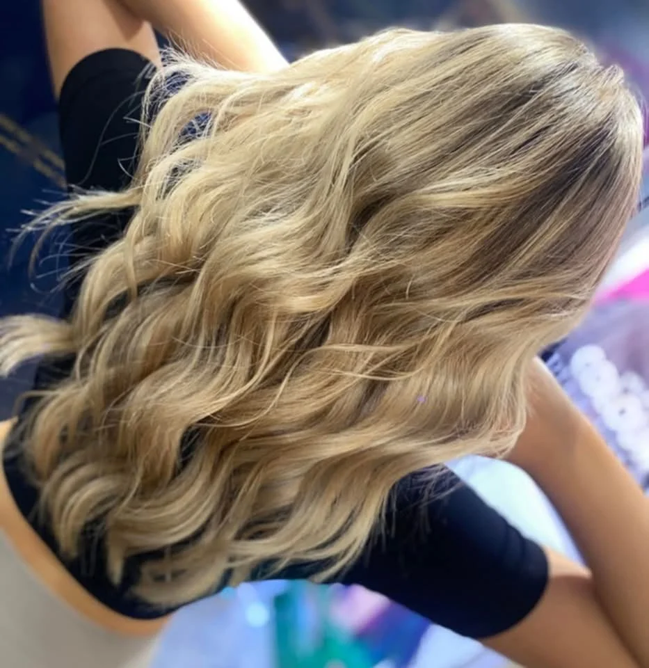 Woman with long blonde wavy hair sitting with her back to the camera, wearing a black top.