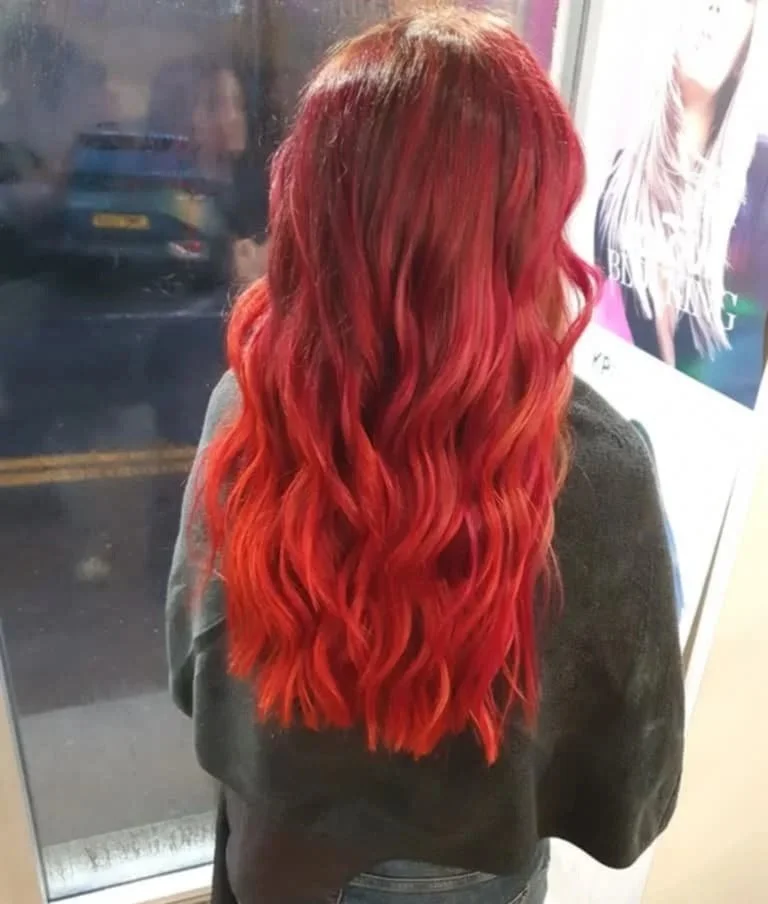 Back view of a woman with long, wavy, vibrant red hair standing in front of a window.