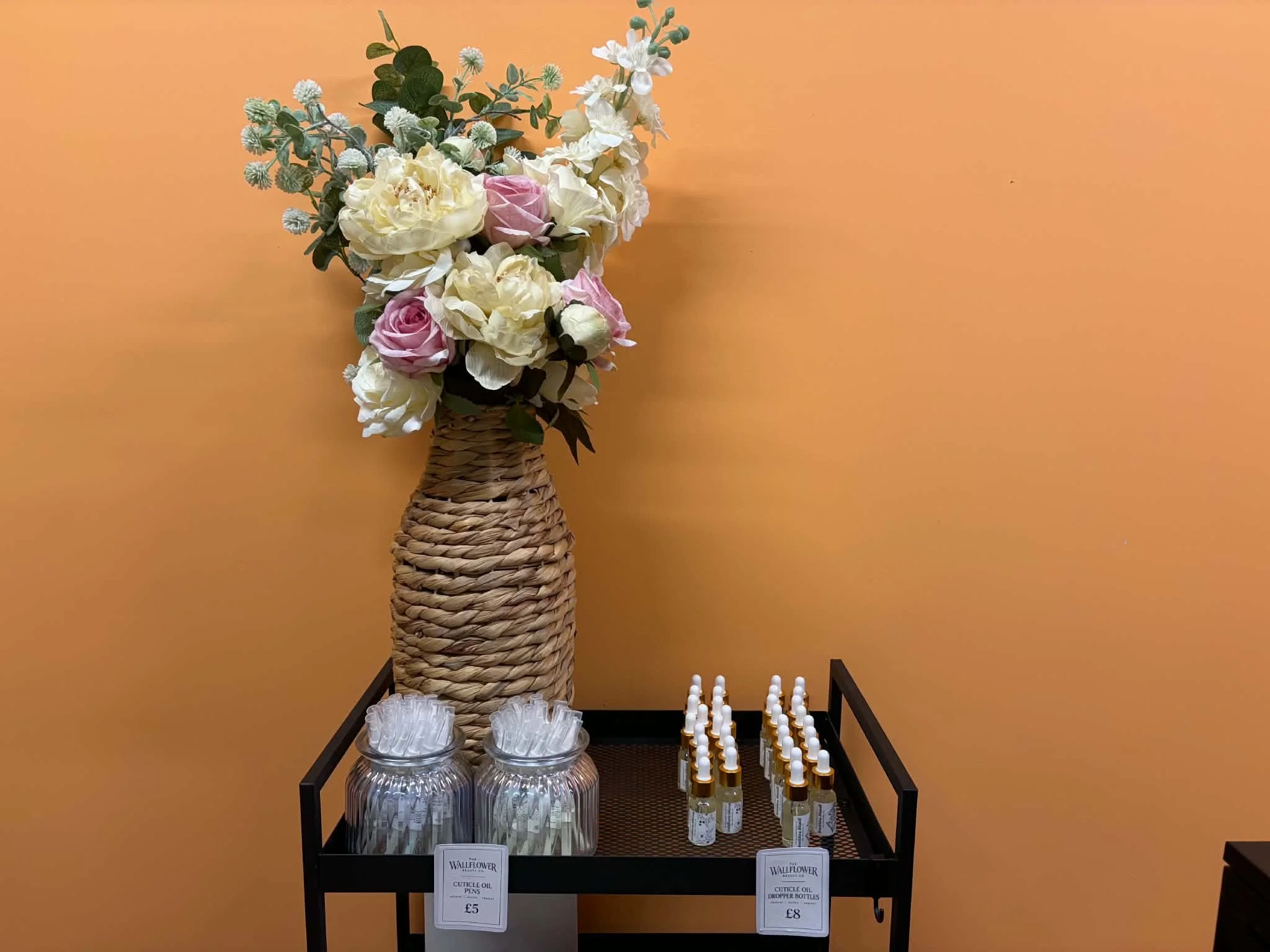 A display tray with candles, dropper bottles of cuticle oil, and tissues, with a large floral arrangement in a woven basket behind it, set against a peach wall.