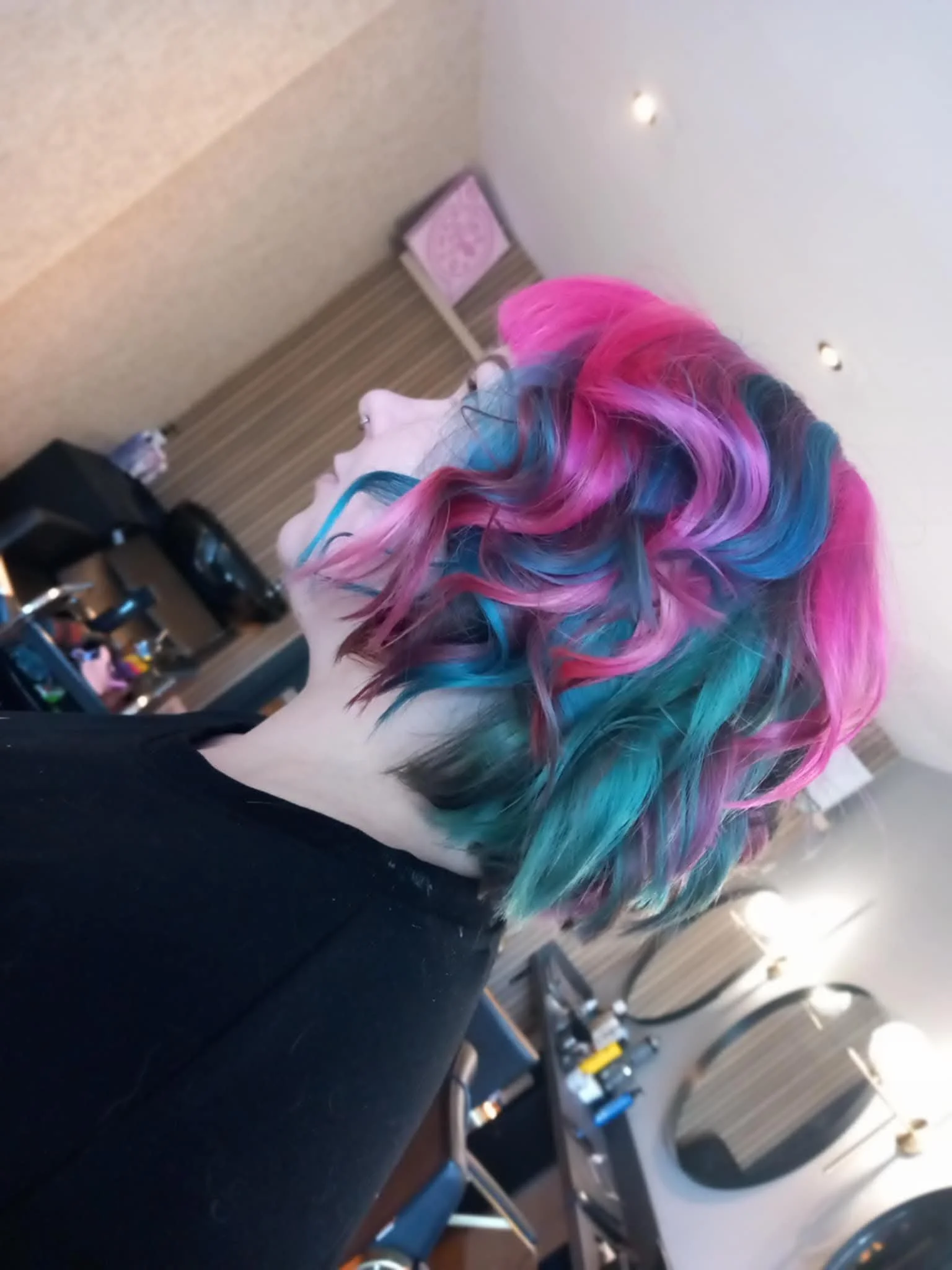 A woman with multicolored, curly hair in shades of pink, purple, and blue. She is seated in a cafe or restaurant, facing sideways, with a side profile visible. The background includes a paper bag, wood-paneled wall, and a washing machine.