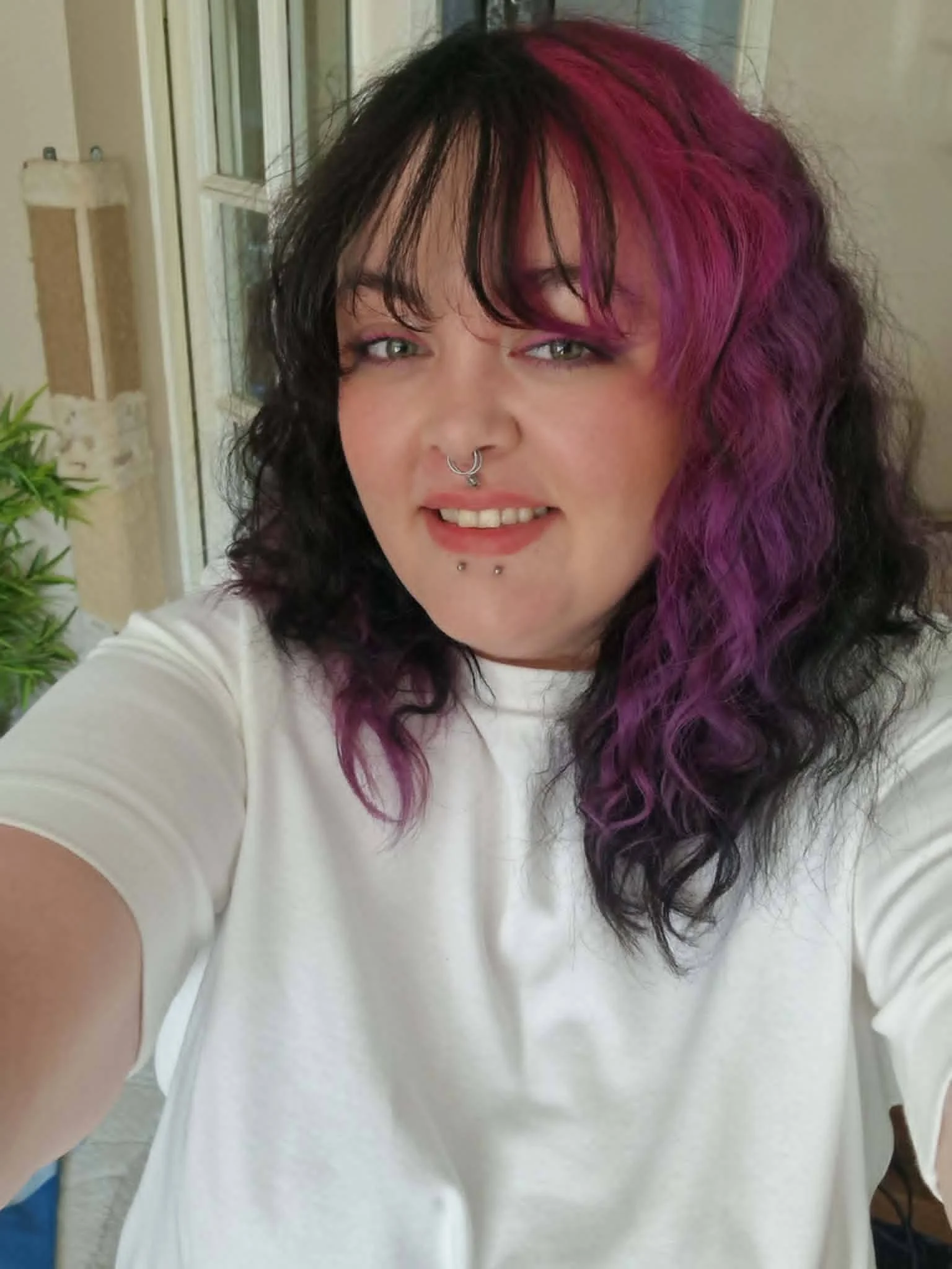 Close-up of a woman with mixed black and purple curly hair, wearing a septum piercing and piercings on her lower lip, smiling slightly, dressed in a white shirt, in front of a blurred outdoor background.
