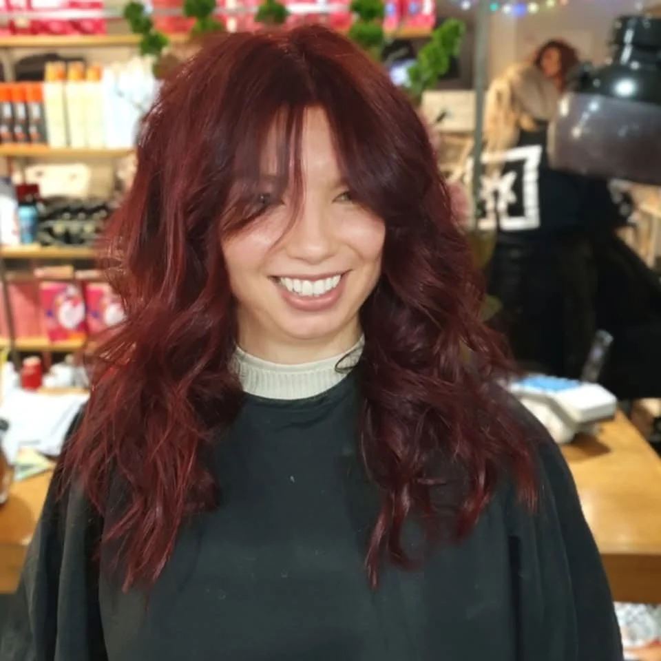 A woman with long, wavy red hair smiling in a cozy setting, likely a café or bookstore, with bookshelves and another person in the background.