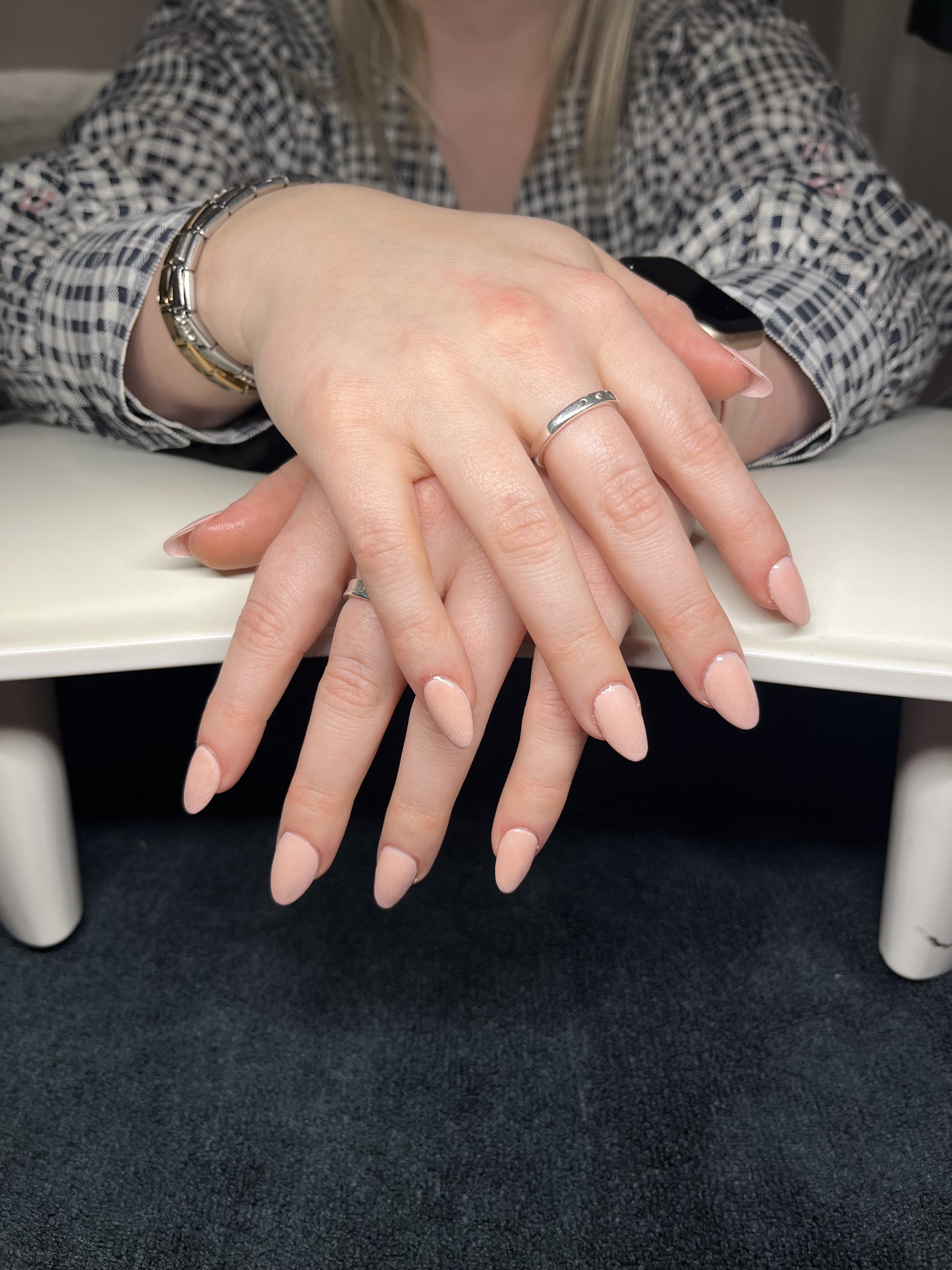 Close-up of a person's hands with manicured nails and silver rings, resting on a black surface. The person is wearing a checkered shirt and a smartwatch on the wrist.