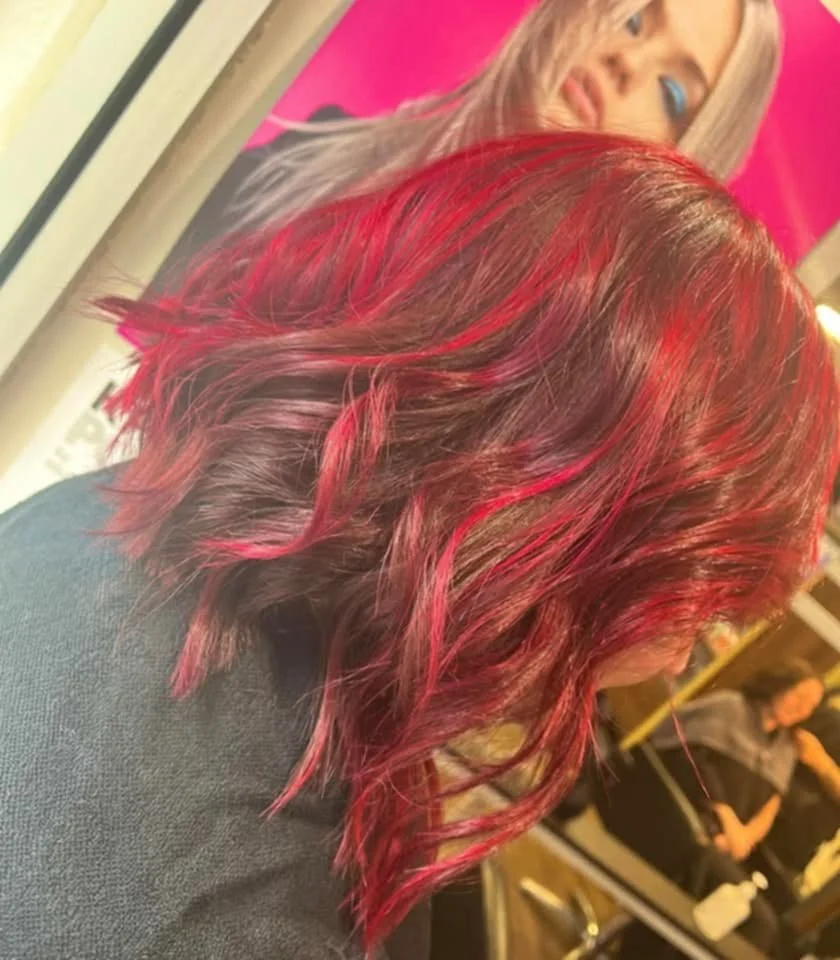 Side view of a woman with wavy red hair at a salon, with a mirror and another person in the background.