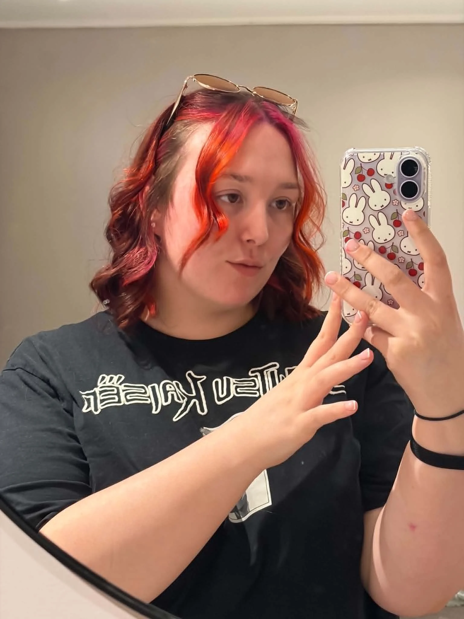A young woman with red and orange wavy hair taking a mirror selfie with her smartphone, which has a bunny pattern case, wearing sunglasses on her head and a black T-shirt.