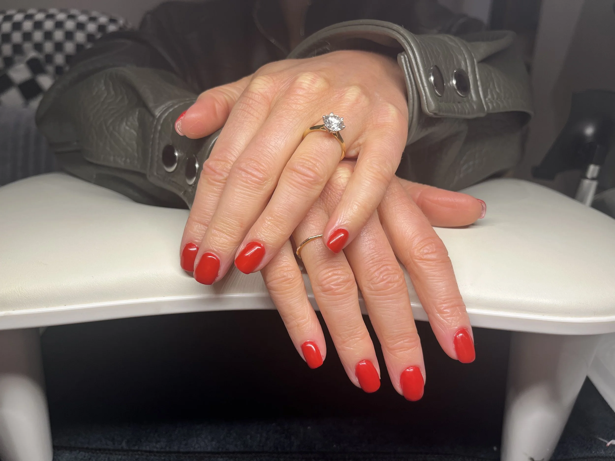 Close-up of a woman's hands with red nail polish, wearing a gold ring with a large diamond, resting on a white surface. A part of her gray sleeve with buttons is visible, and the background includes a black and white checkered pattern.