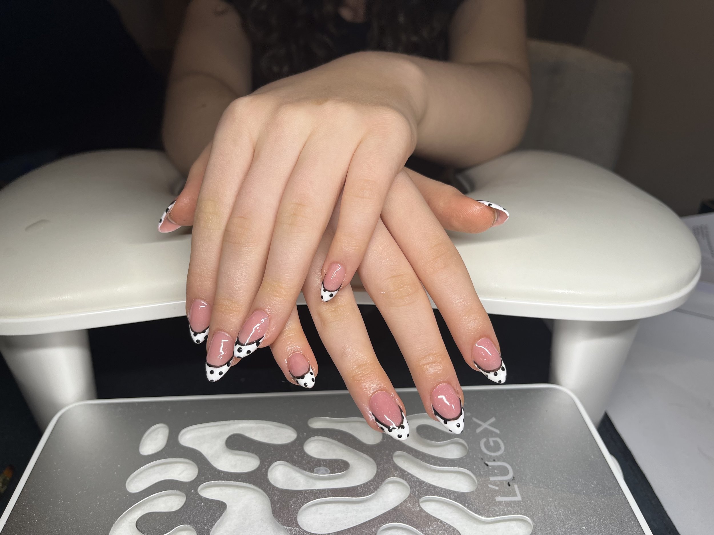 Hands with manicured nails resting on a white surface, with a nail wrap or design featuring black and white polka dots and black trim. In the background, a person's torso and arms are partially visible, wearing a dark top with lace details, and they are seated at a white table or nail station.