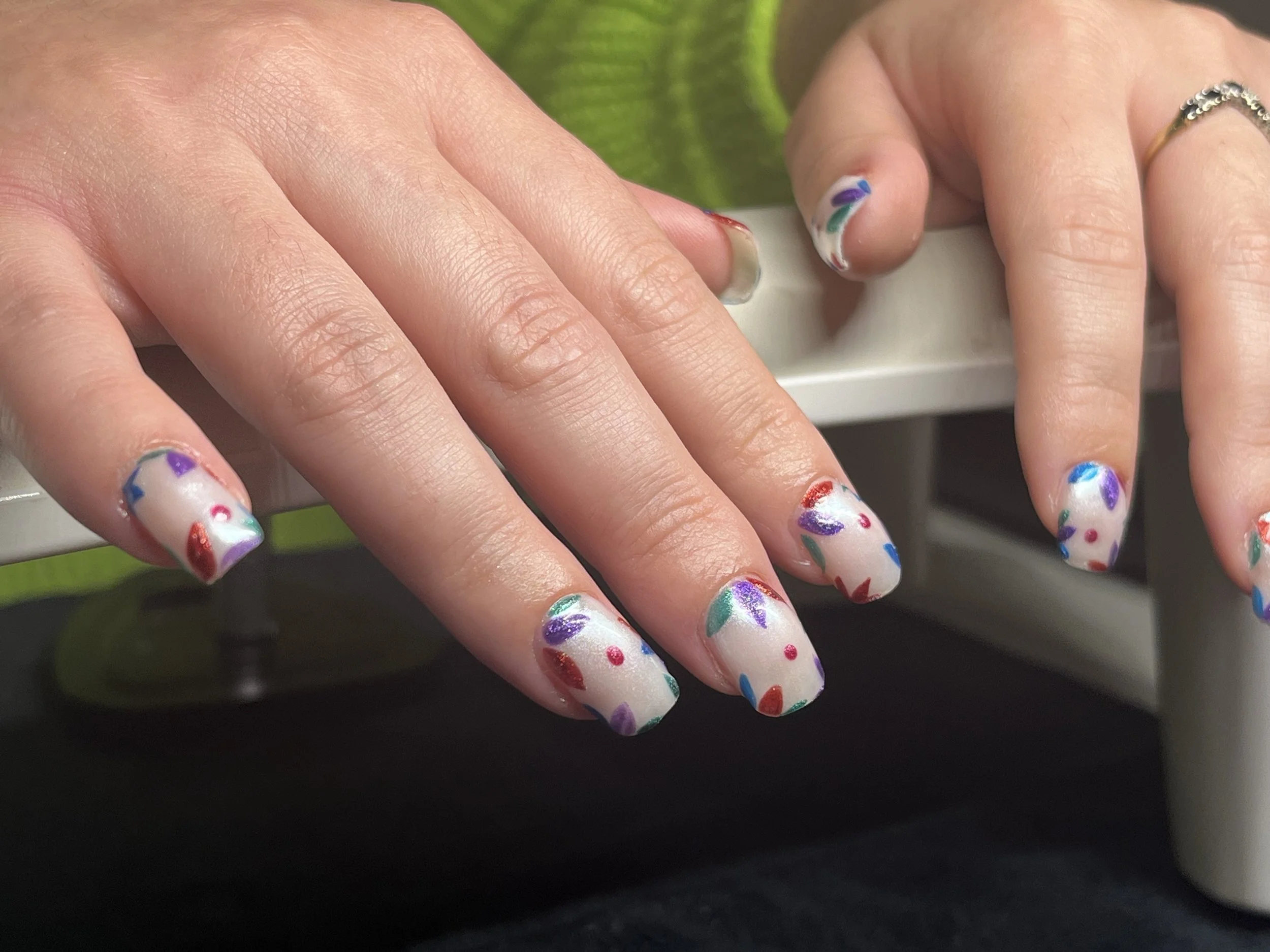 Close-up of a person's hand with colorful, patterned nails, and a silver ring on the finger. The background includes a green leaf.