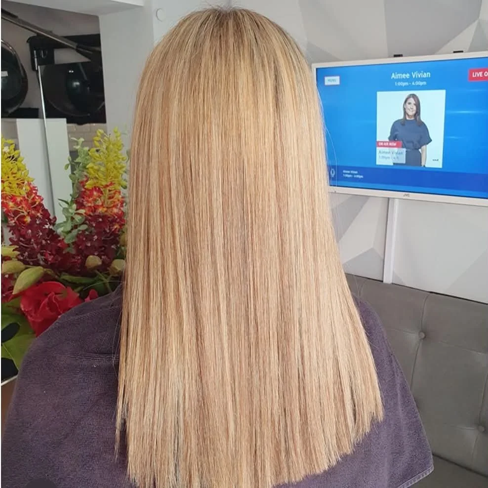 Back view of a woman with long, straight, blonde hair sitting in a salon or waiting area, with a bouquet of colorful flowers beside her and a monitor screen displaying a woman named Aimee Vivian.