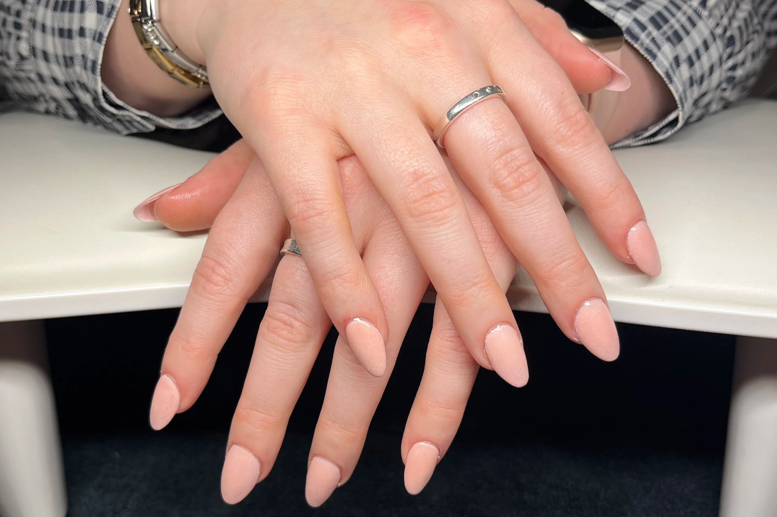 Close-up of a person's hands resting on a white surface, with one hand on top of the other. The person is wearing rings on their fingers and has neatly manicured nails with a natural pink color. They are dressed in a black and white patterned shirt with a silver bracelet visible on the wrist.
