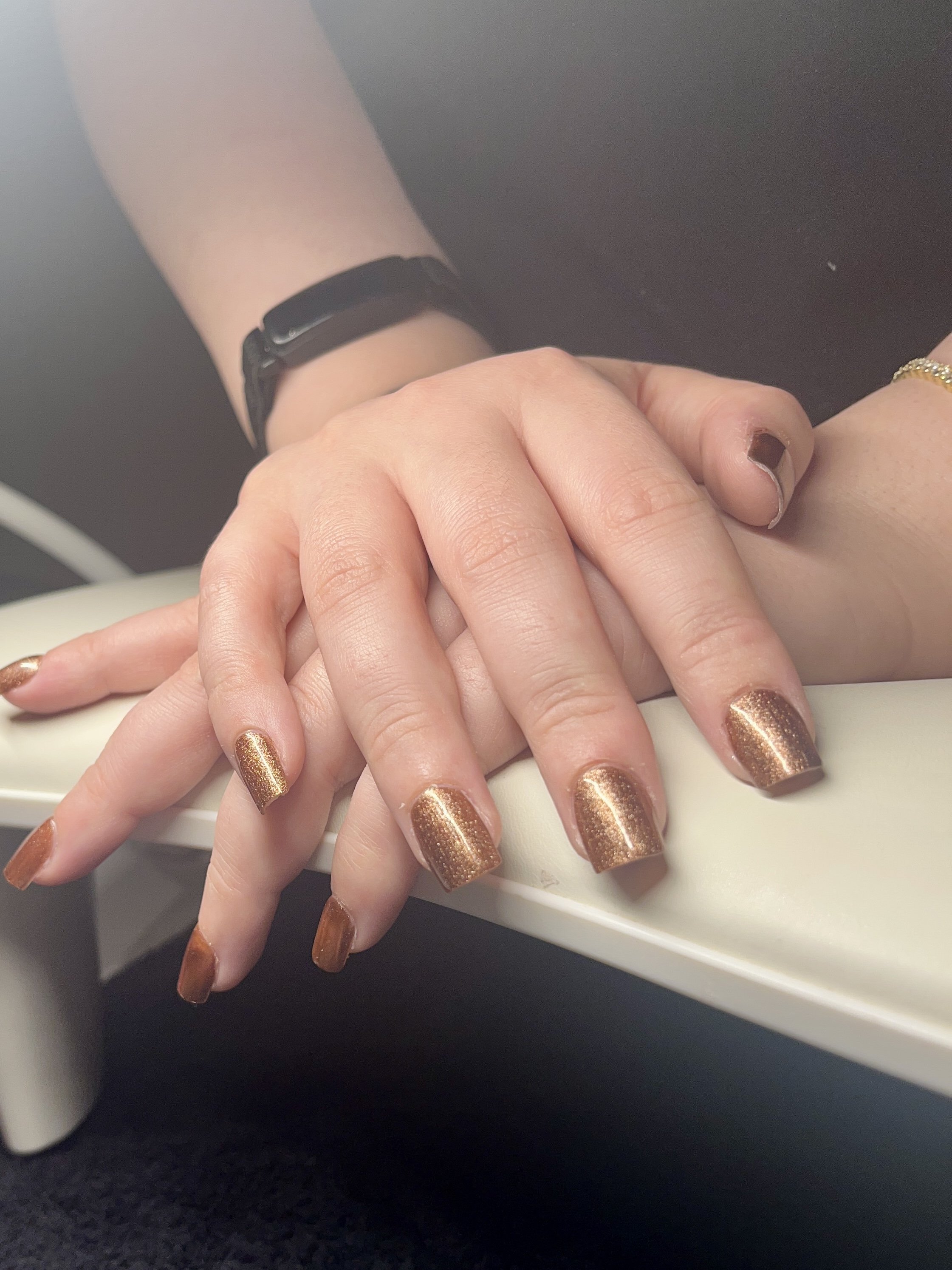 Close-up of hands with gold-colored, glittery nail polish on fingernails, resting on a white surface, with a black background and part of a person's wrist wearing a black watch visible.