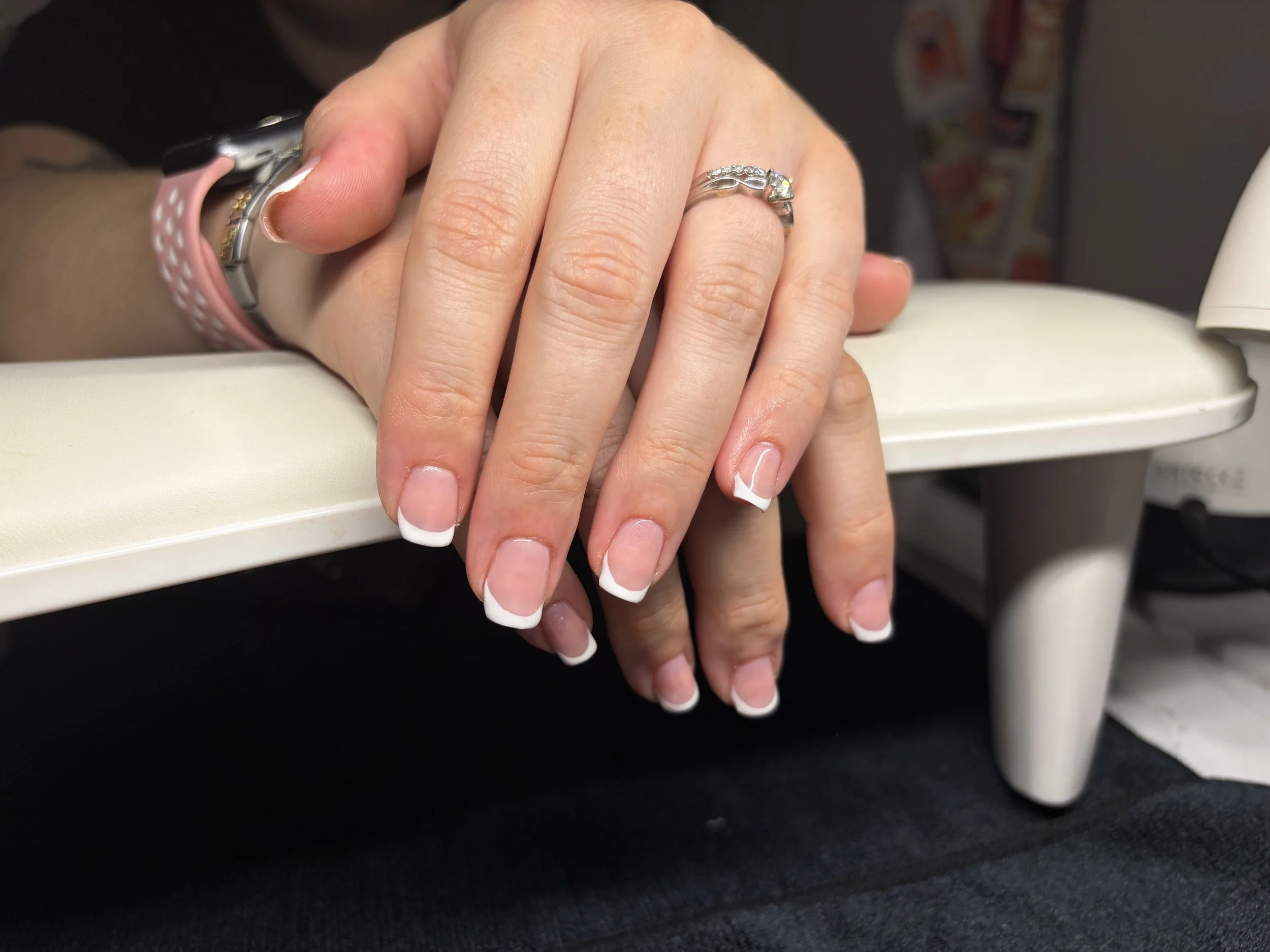 Close-up of a person's hand resting on a white surface, showing manicured nails with French tips, wearing a silver ring with a large clear gemstone and smaller stones. The person has a pink watch with a black face on their wrist, and the background contains a blurred wall with colorful items.