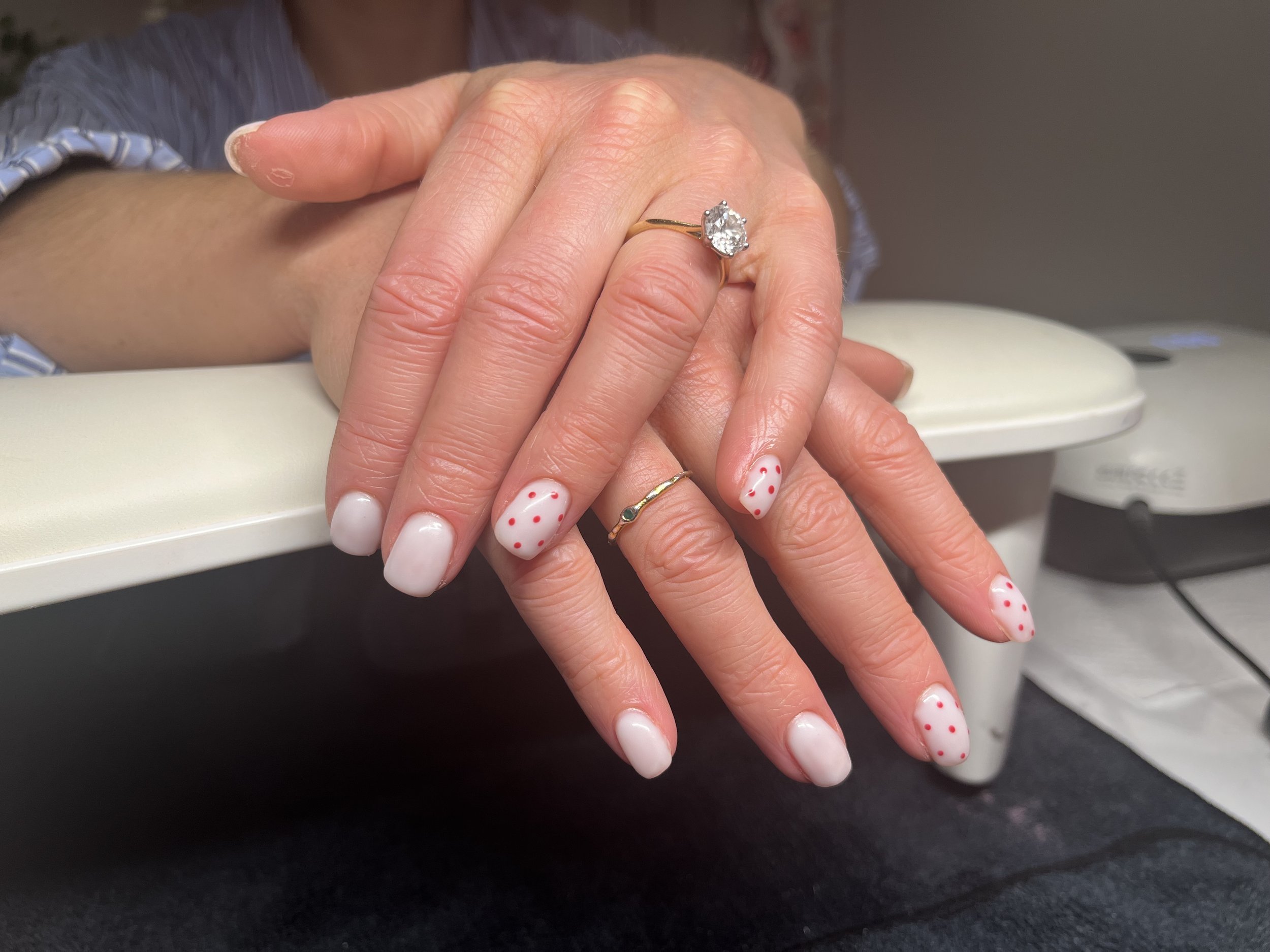 Close-up of a woman’s hand with a diamond engagement ring on her ring finger, resting on a surface, with her nails painted white with red polka dots, and dressed in a blue and white striped shirt.