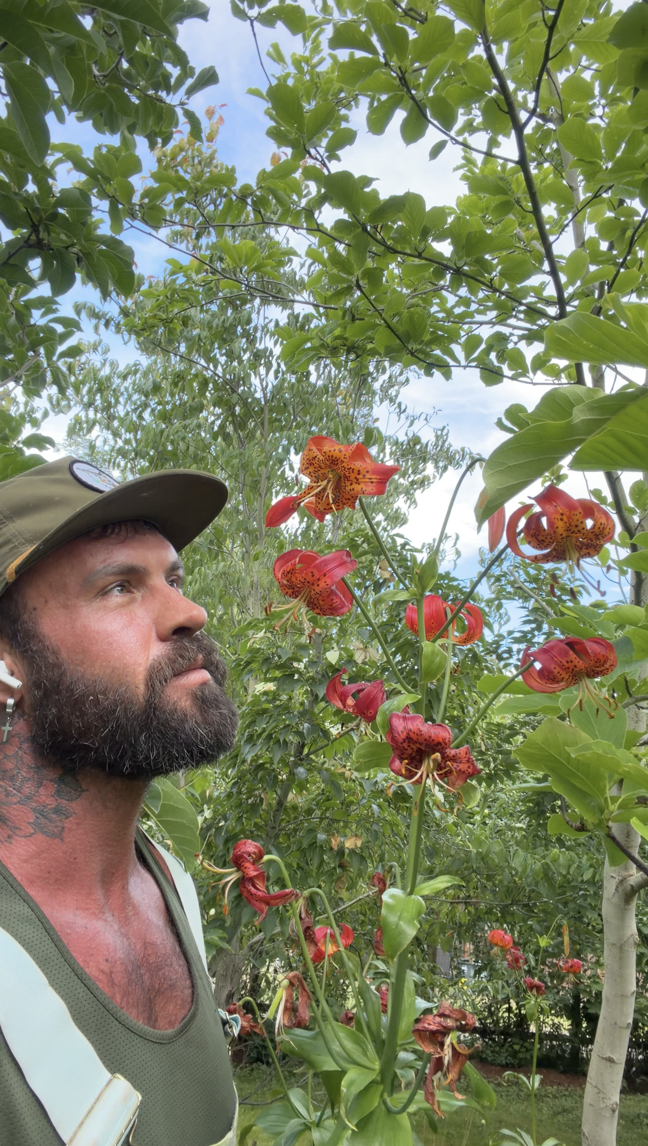 A man with a beard and tattooed chest wearing a green tank top and a hat, standing outdoors among green trees and red-orange lilies with some withered petals and some in bloom.