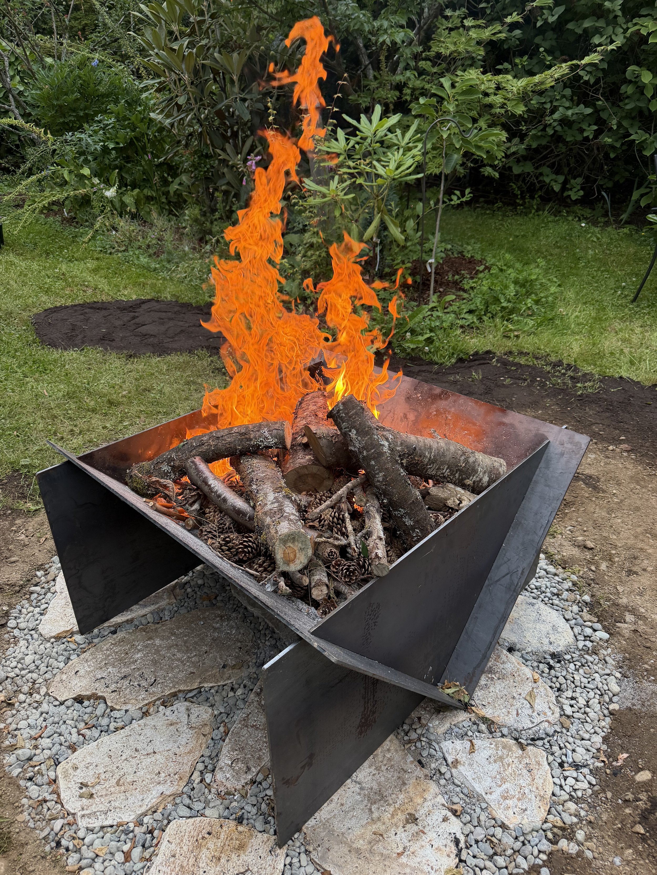 Outdoor fire pit with logs and flames in a garden with green plants and trees.