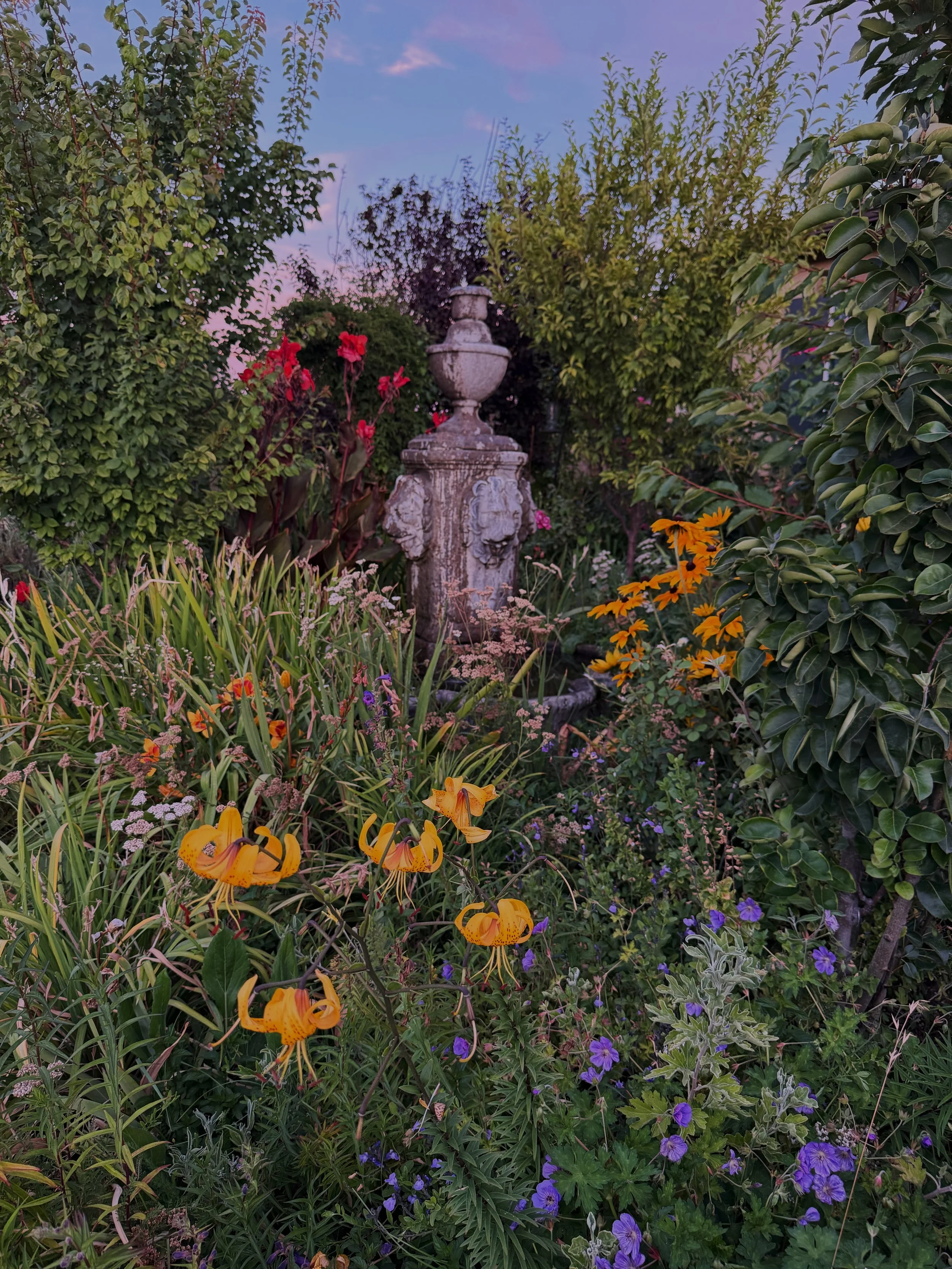 A garden scene with a stone fountain surrounded by various colorful flowers and lush green plants, under a pink and purple sky.