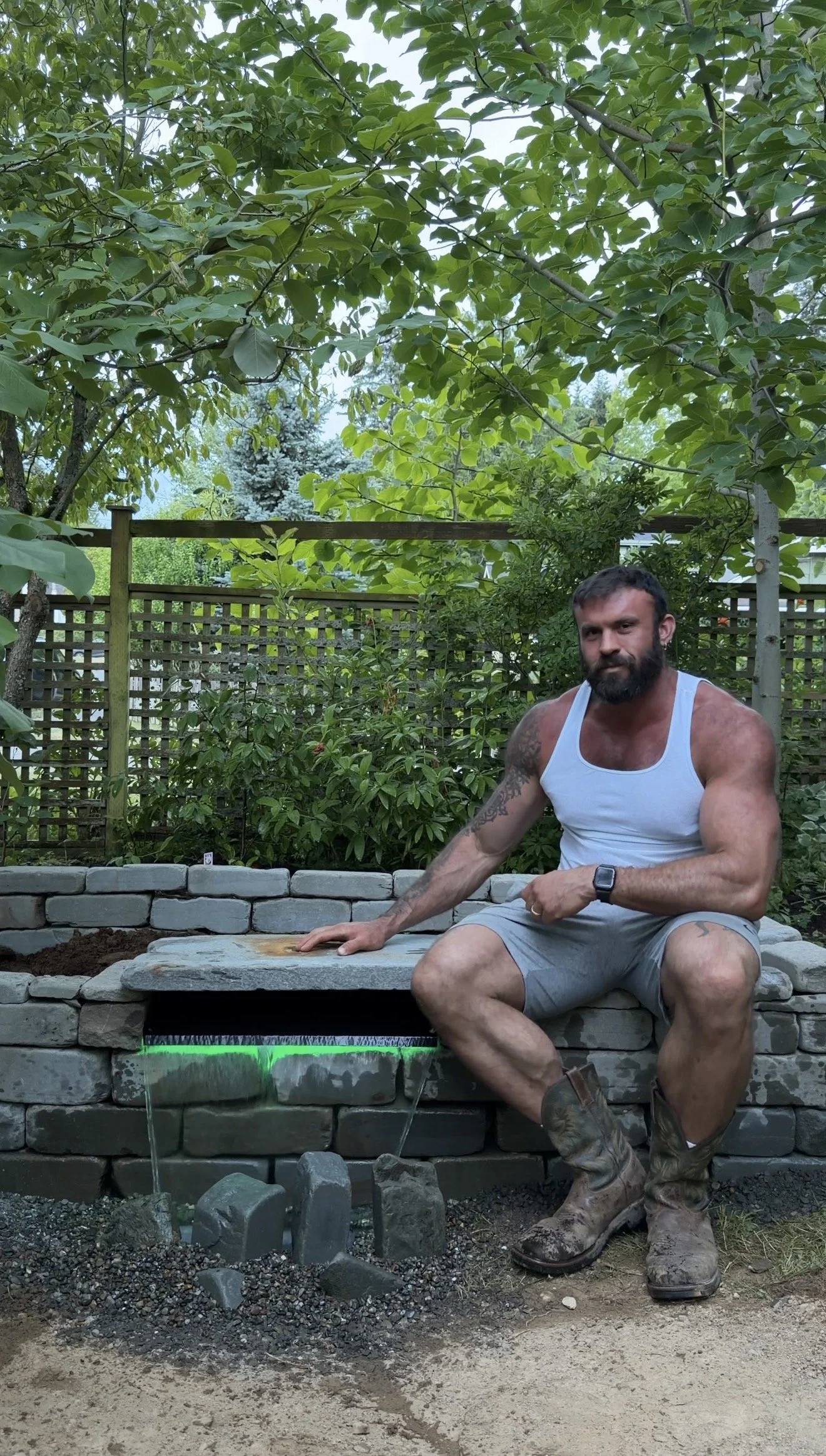 A muscular man with a beard, wearing a white tank top, gray shorts, and camouflage boots, sitting on a stone structure in a backyard garden with green trees and a wooden fence in the background.