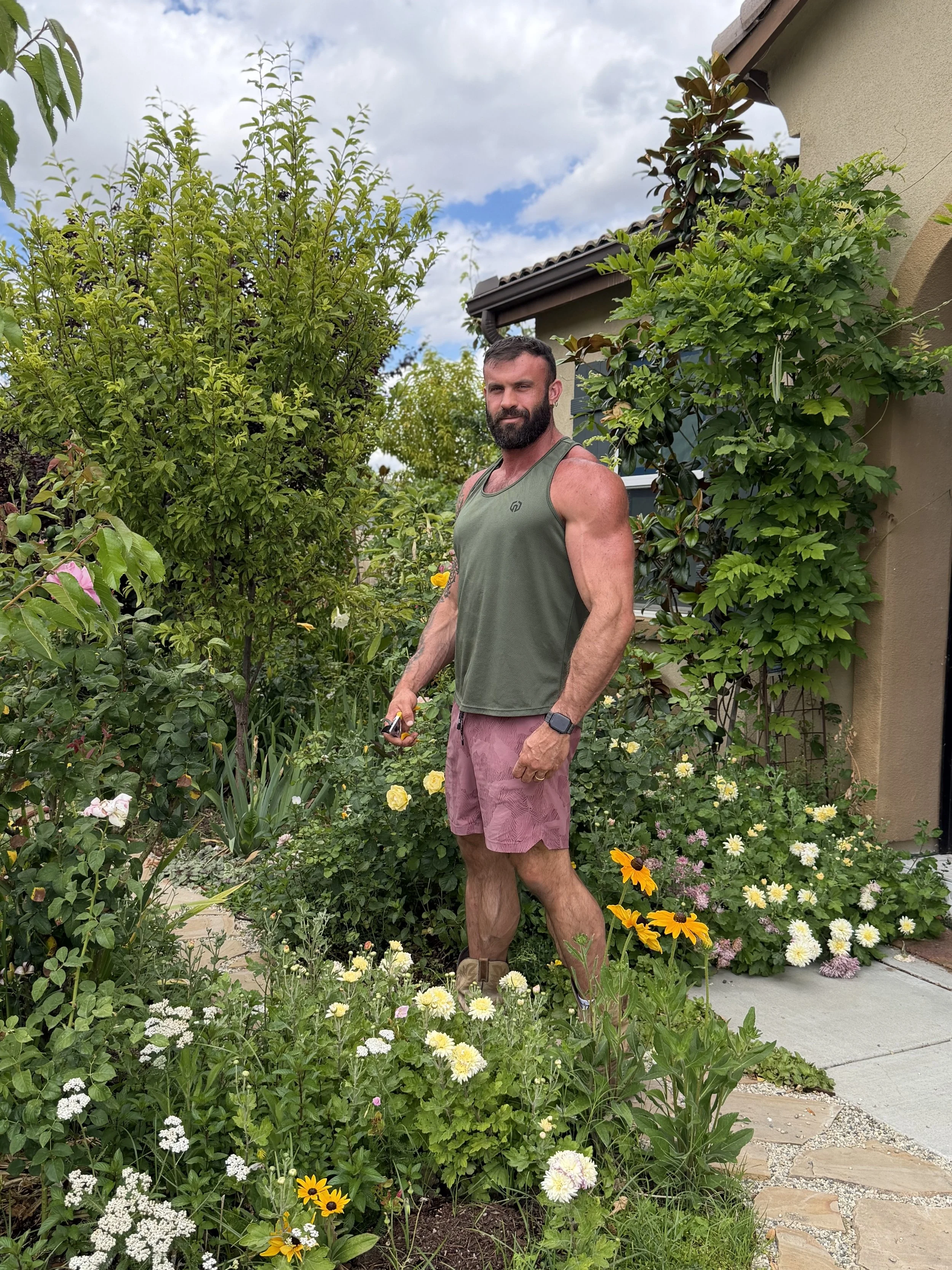 A muscular man with a beard, wearing a green sleeveless shirt and pink shorts, stands in a garden filled with various flowers and greenery. He is holding a garden tool in his right hand and looking at the camera with a slight smile.