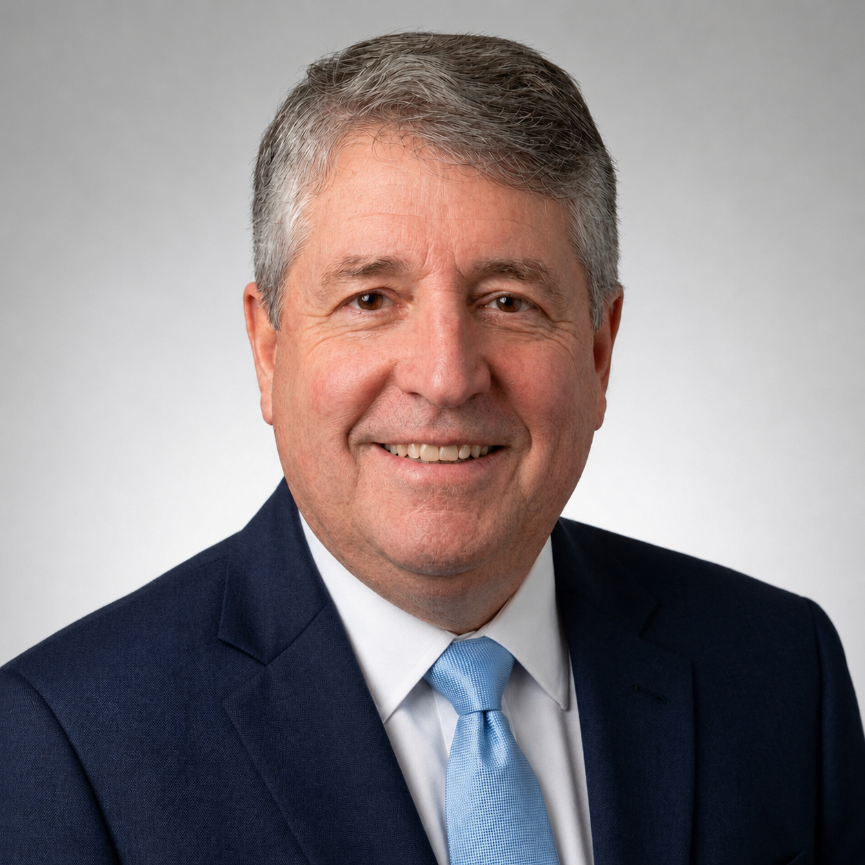 Professional headshot of a middle-aged man with gray hair, wearing a navy suit, white shirt, and light blue tie, smiling against a plain gray background.