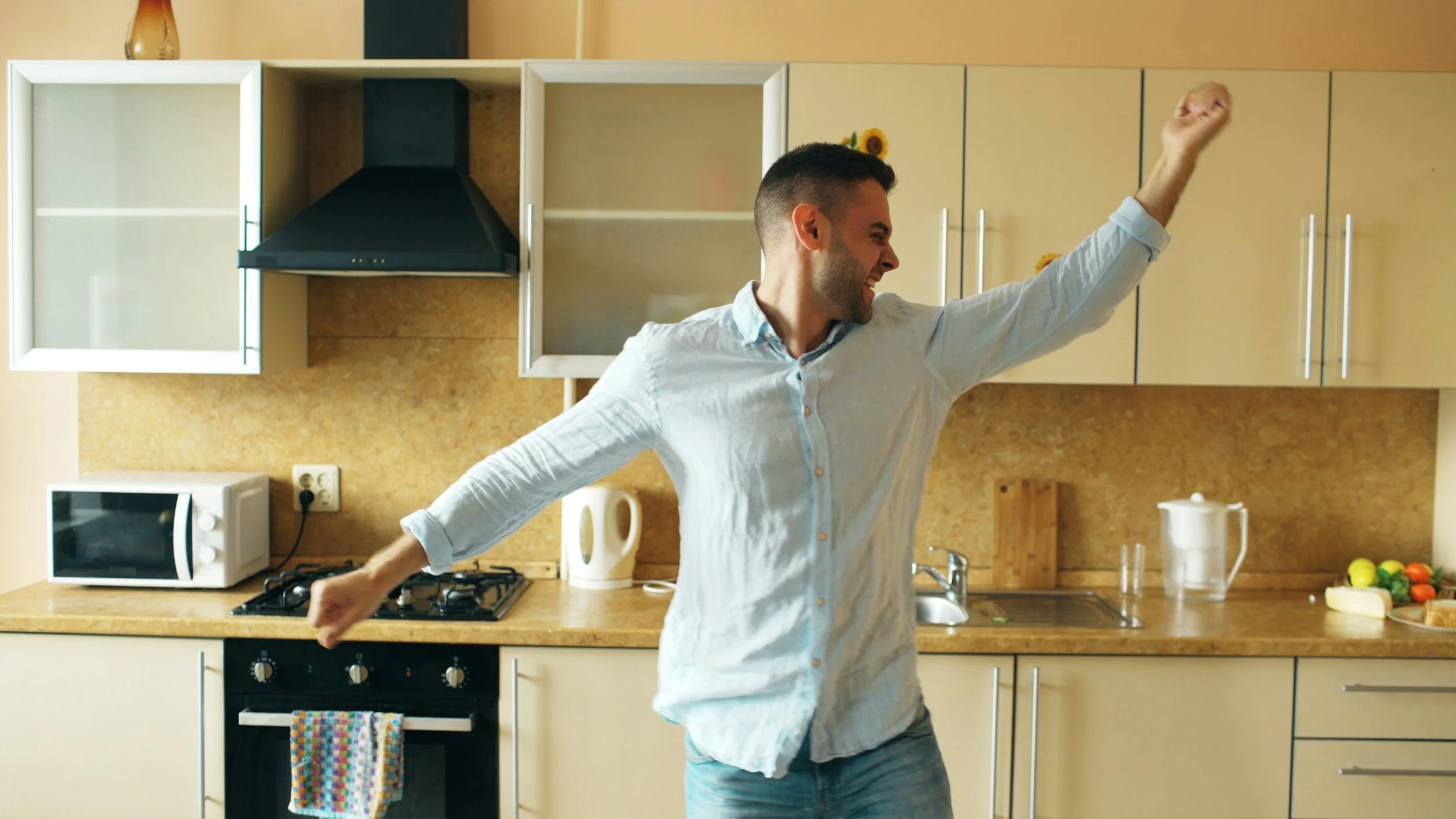 A man dancing or celebrating in a kitchen with beige cabinets, a black range hood, a microwave, and various kitchen items on the countertop.