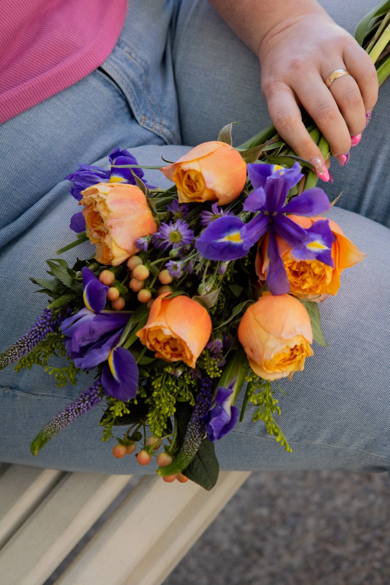 A person sitting on a grey bench holding a bouquet of orange, purple, and pink flowers, wearing light blue jeans and a pink shirt.