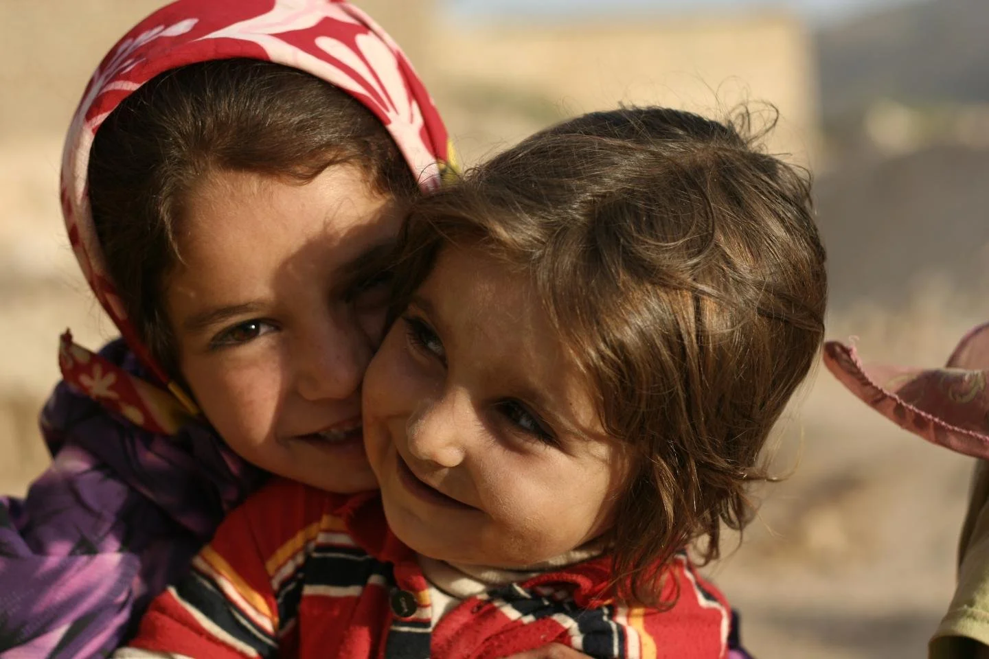 Two young girls hugging and smiling outdoors, one wearing a red and white headscarf, the other with brown hair.