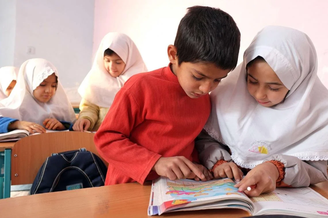 A teacher helping a young student with a colorful book in a classroom, with other children in headscarves in the background.