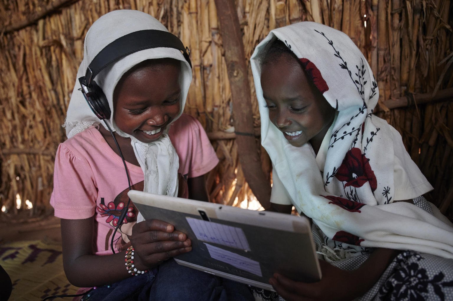 Two young African girls sit inside a wooden hut, smiling and looking at a tablet device. One girl is wearing a pink shirt and headphones, and the other is wearing a light-colored headscarf with floral patterns.