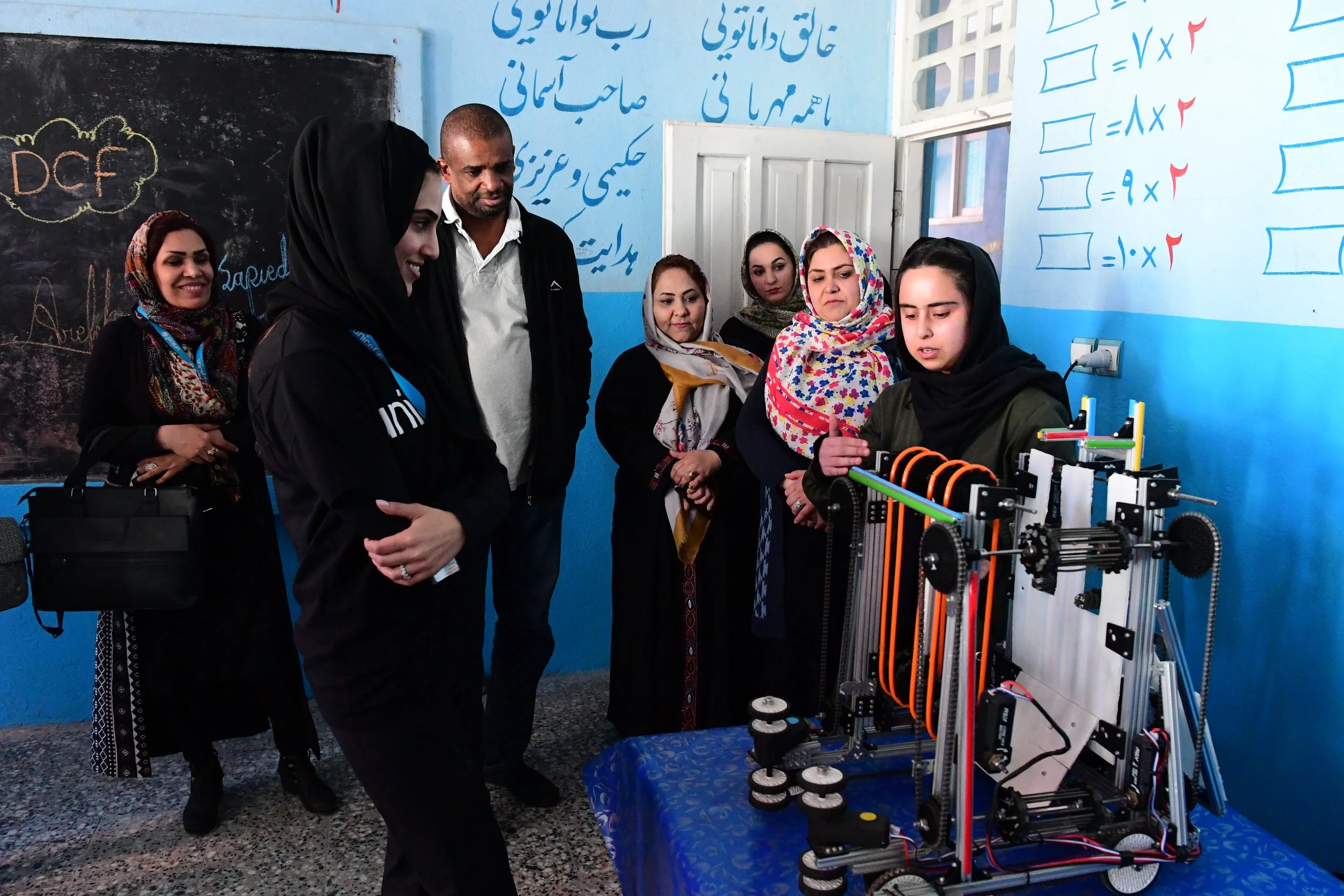 Group of women and a man observing a robotic machine at an indoor educational event, with colorful writings on blue walls.