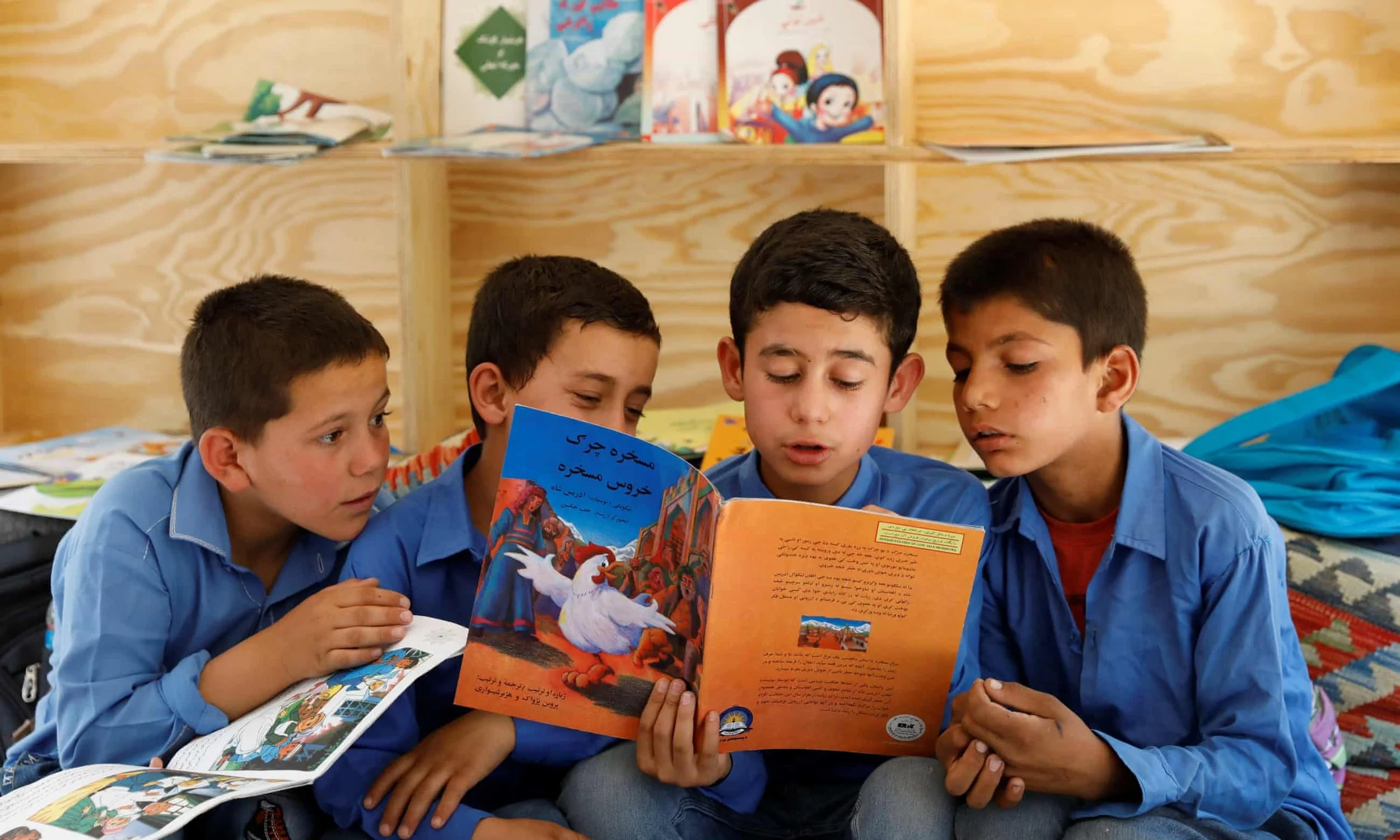 Four young boys sitting together on a colorful rug in a wooden room, reading a children's book with vibrant illustrations. The boys are wearing blue shirts and appear engaged in the story.
