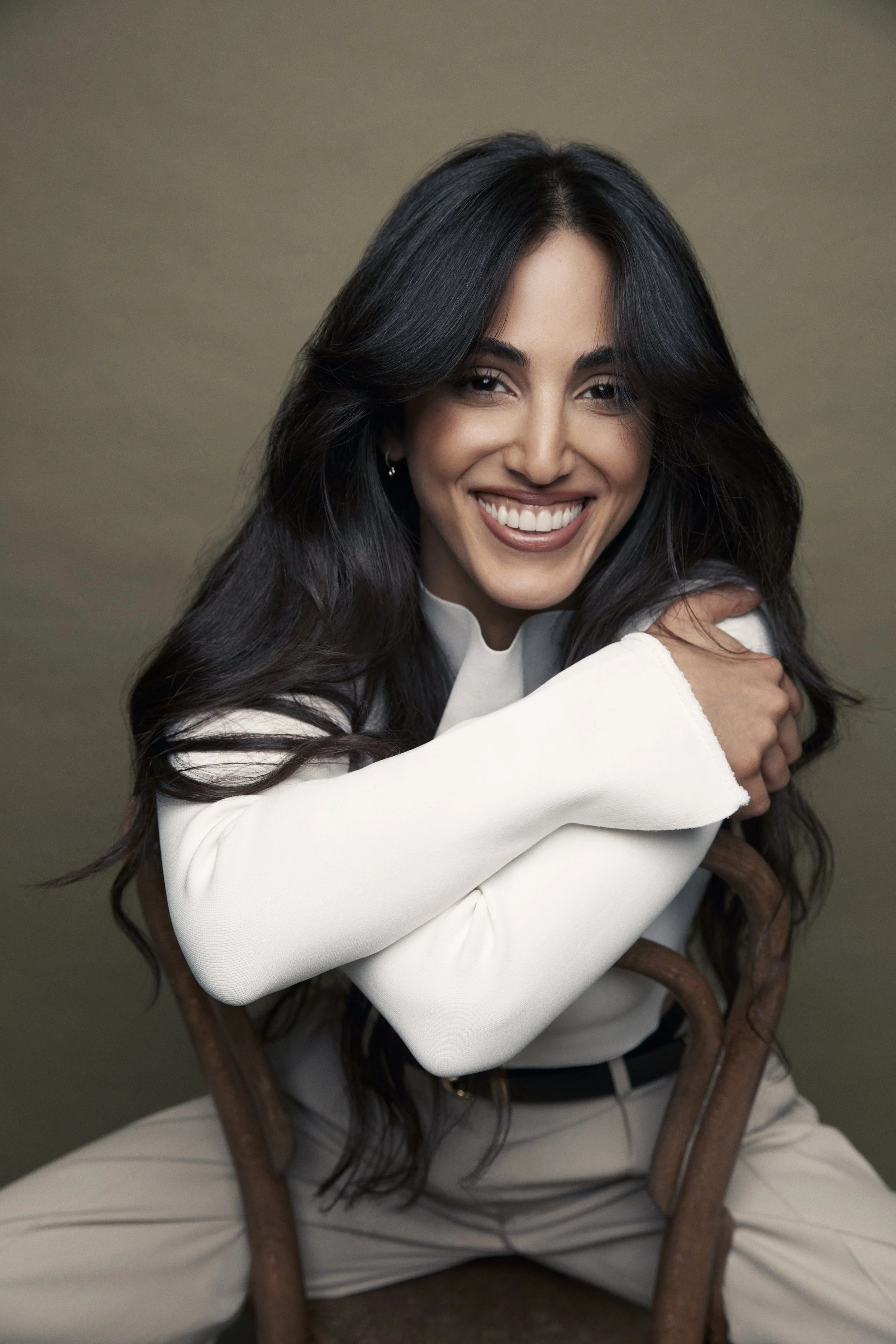 A smiling woman with long dark hair, wearing a white top and beige pants, sitting on a wooden chair with a gray background.