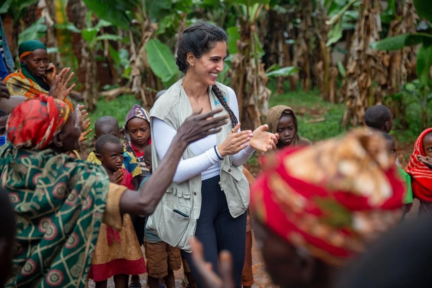 A woman with dark hair in a braid smiling among local villagers with children and women, in a rural setting with banana trees in the background.