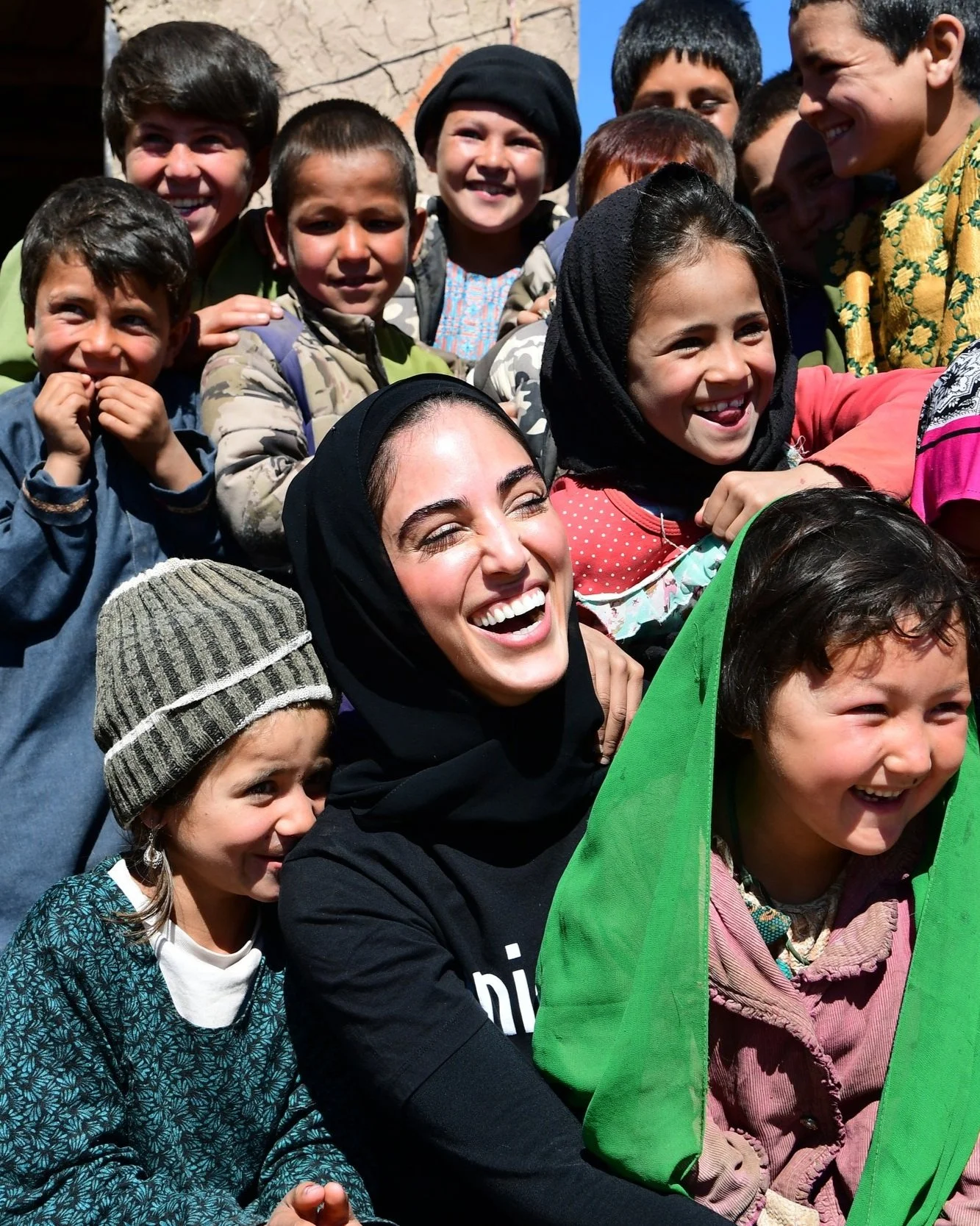 A diverse group of children and a woman, smiling and laughing, outside on a sunny day, standing close together.
