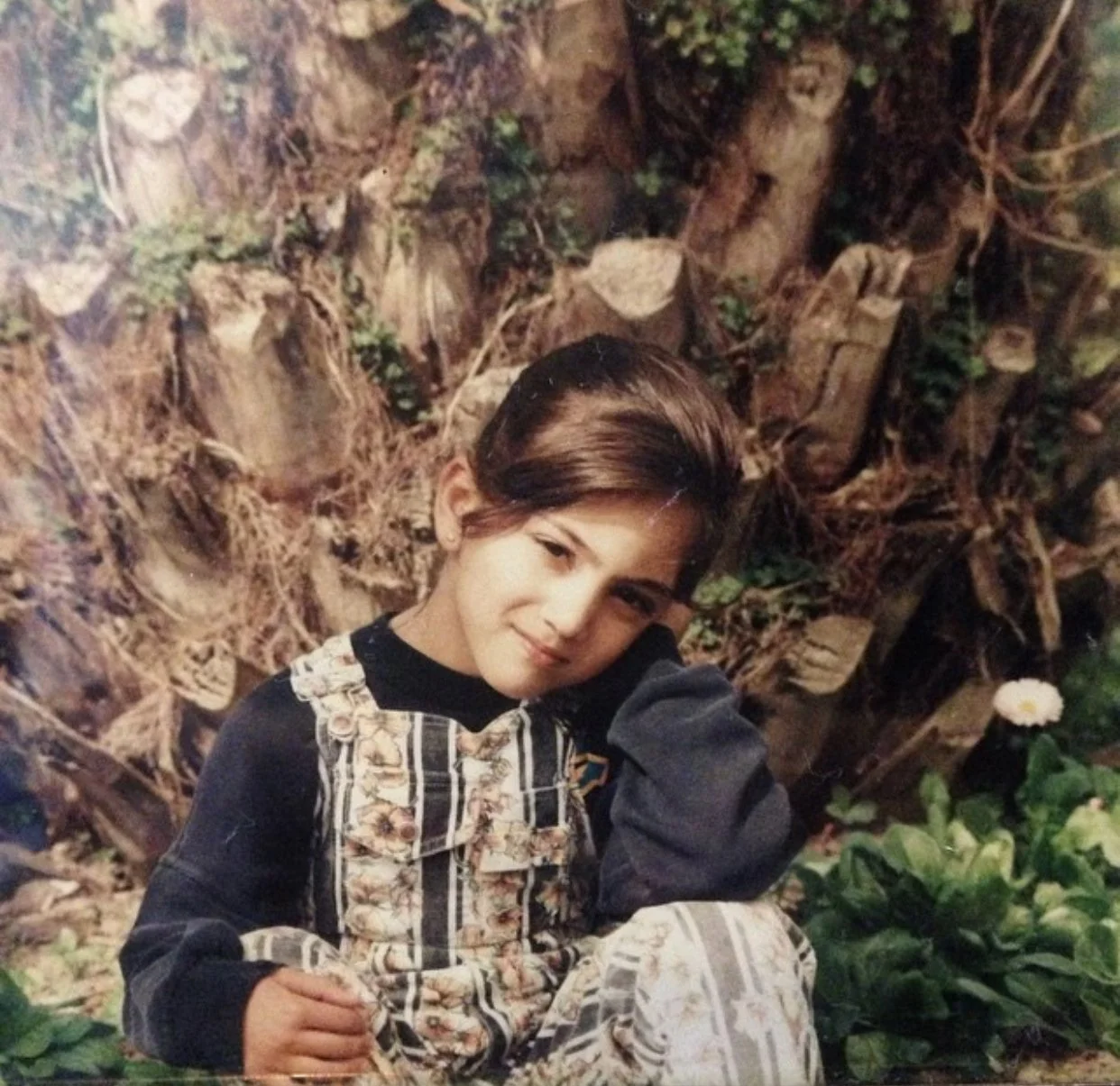A young girl with short, dark hair smiling and resting her head on her hand, seated outdoors against a background of a dry, rocky area with some green plants and a single white flower.