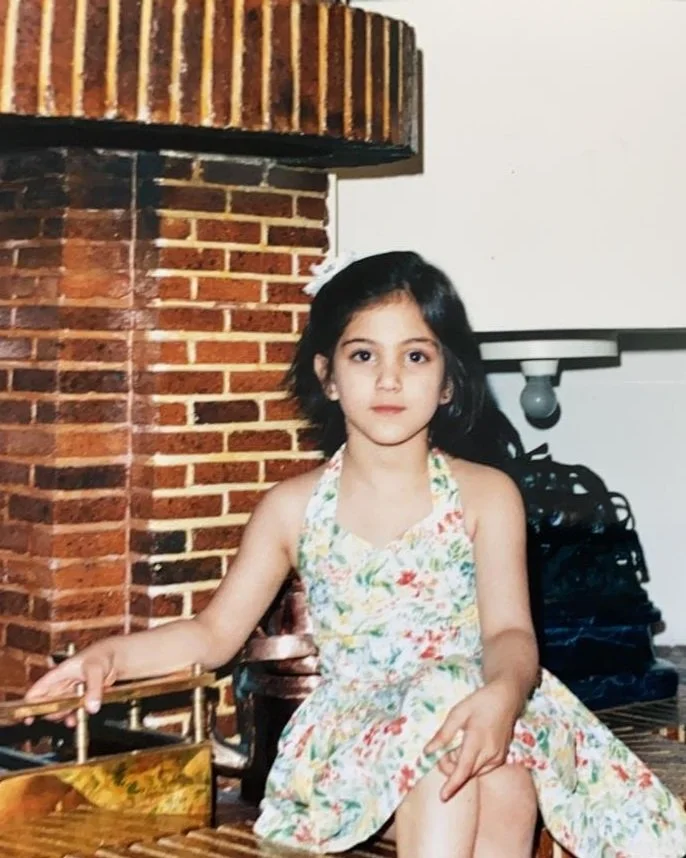 A young girl with dark hair sitting on a wooden chair in front of a brick wall, wearing a floral dress, looking at the camera.