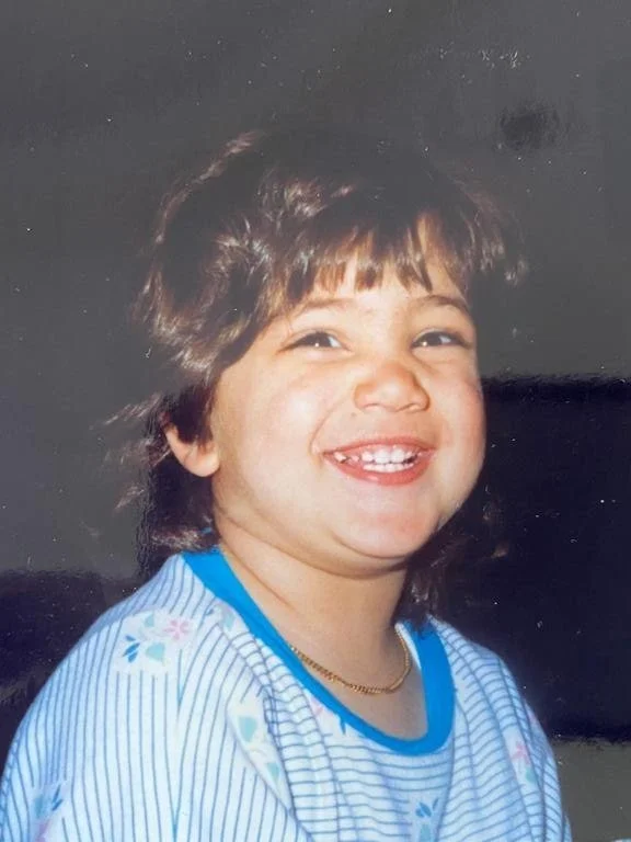 Smiling young child with short brown hair wearing a blue-striped shirt with floral pattern and a gold necklace.