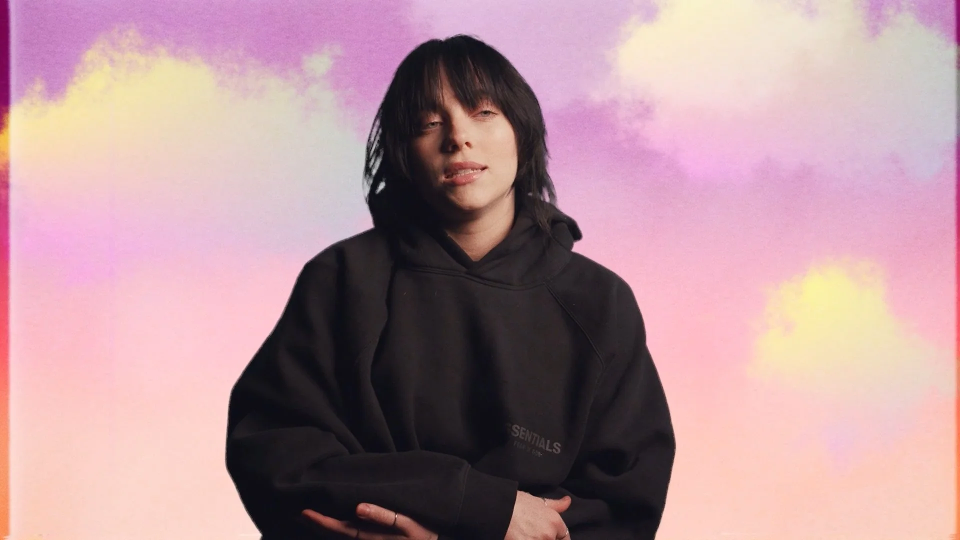 A person with black hair and a black hoodie sitting against a pastel-colored sky background with clouds.