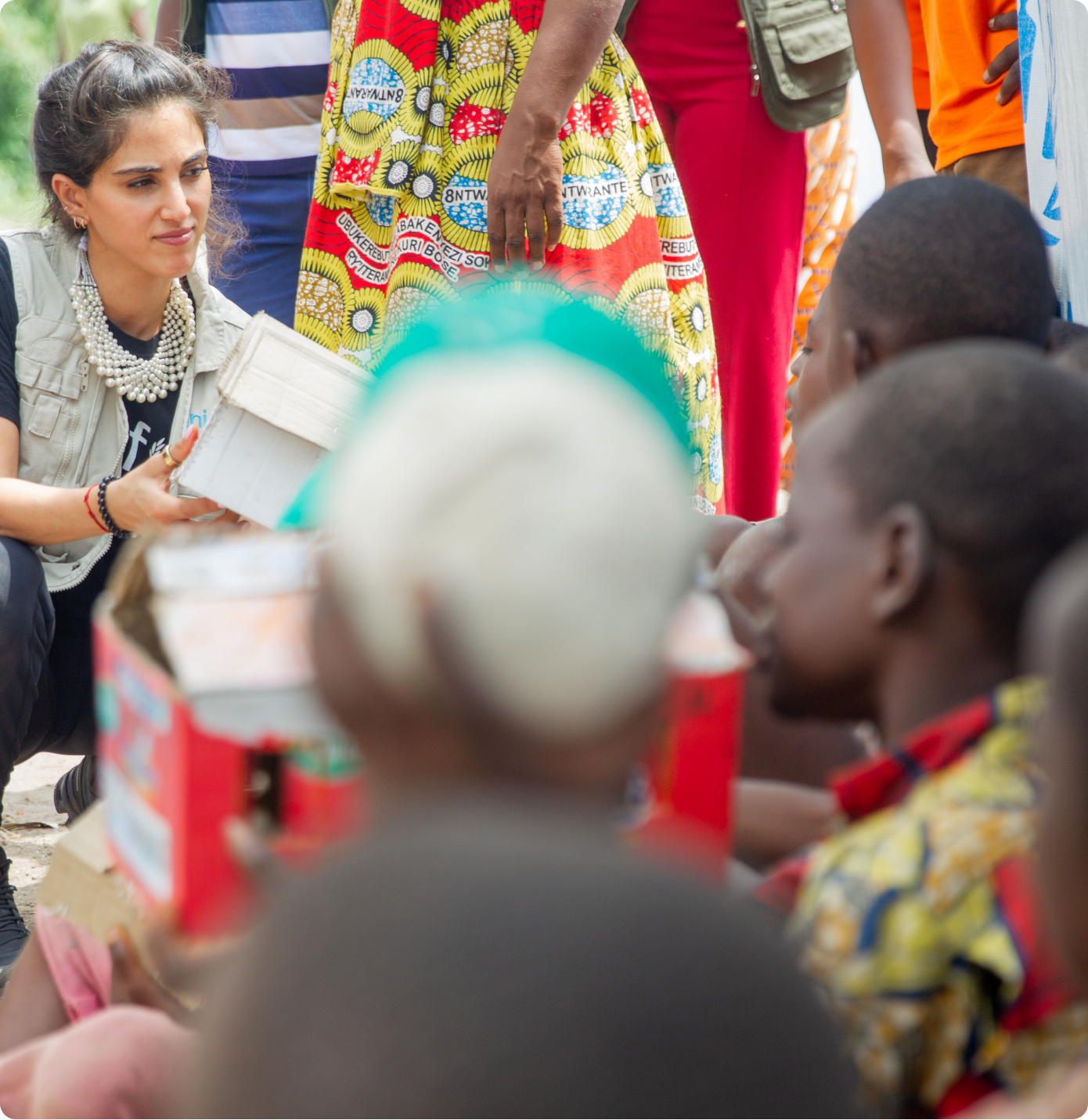 A woman with dark hair and a pearl necklace sitting on the ground, holding a box, engaged with a group of children sitting around her, with a person in colorful clothing standing nearby.