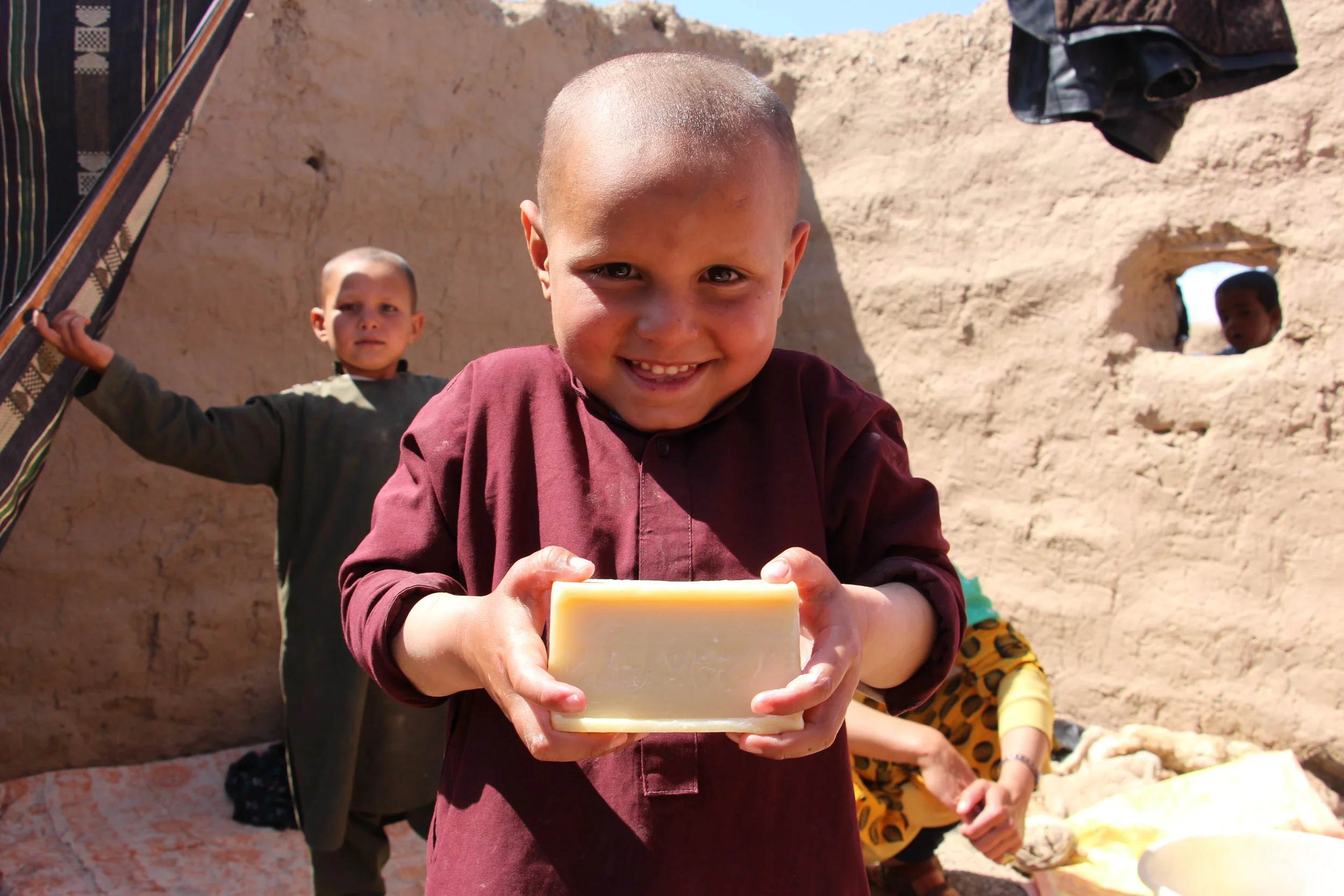 A young boy with a shaved head smiling and holding a bar of soap outdoors, with other children and a mud wall in the background.