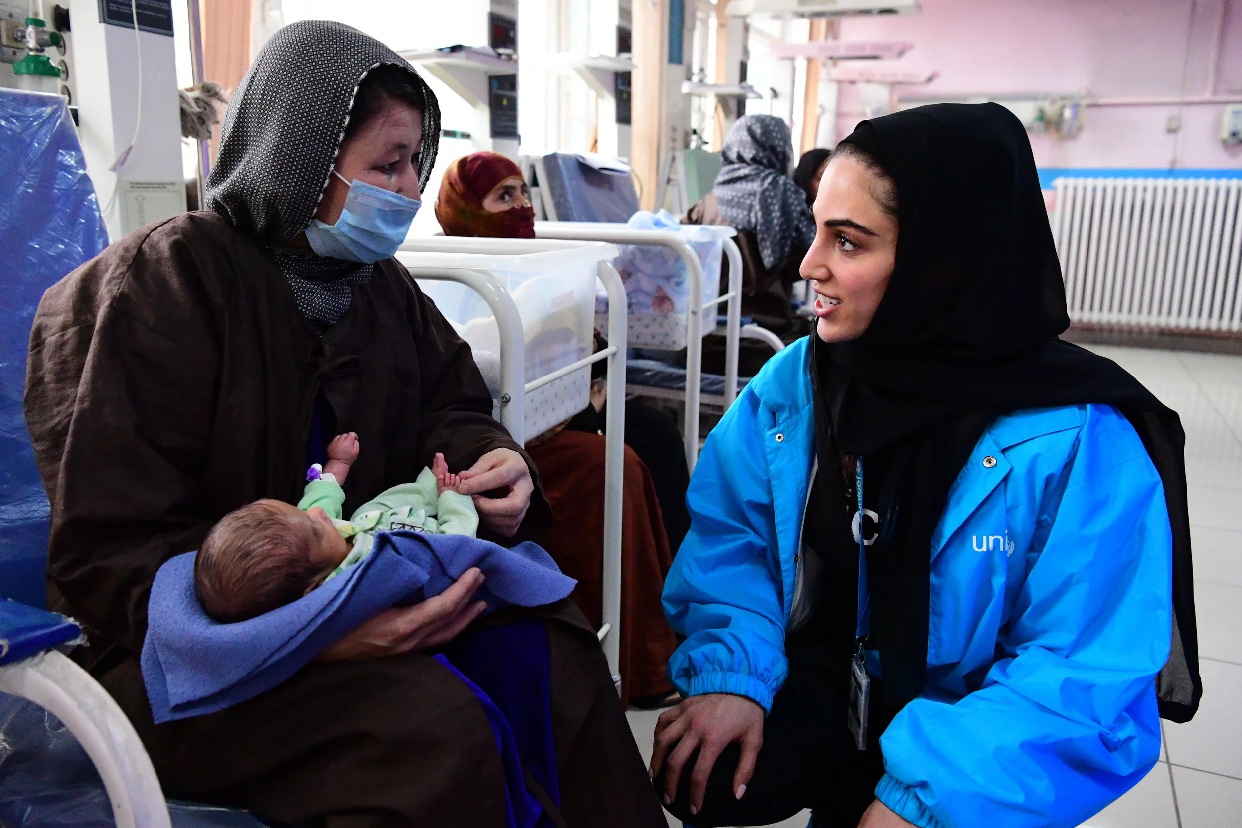 A healthcare worker in blue uniform is talking to a woman wearing a face mask and headscarf, while holding a newborn baby wrapped in a blanket, in a hospital or clinic setting with other patients in the background.