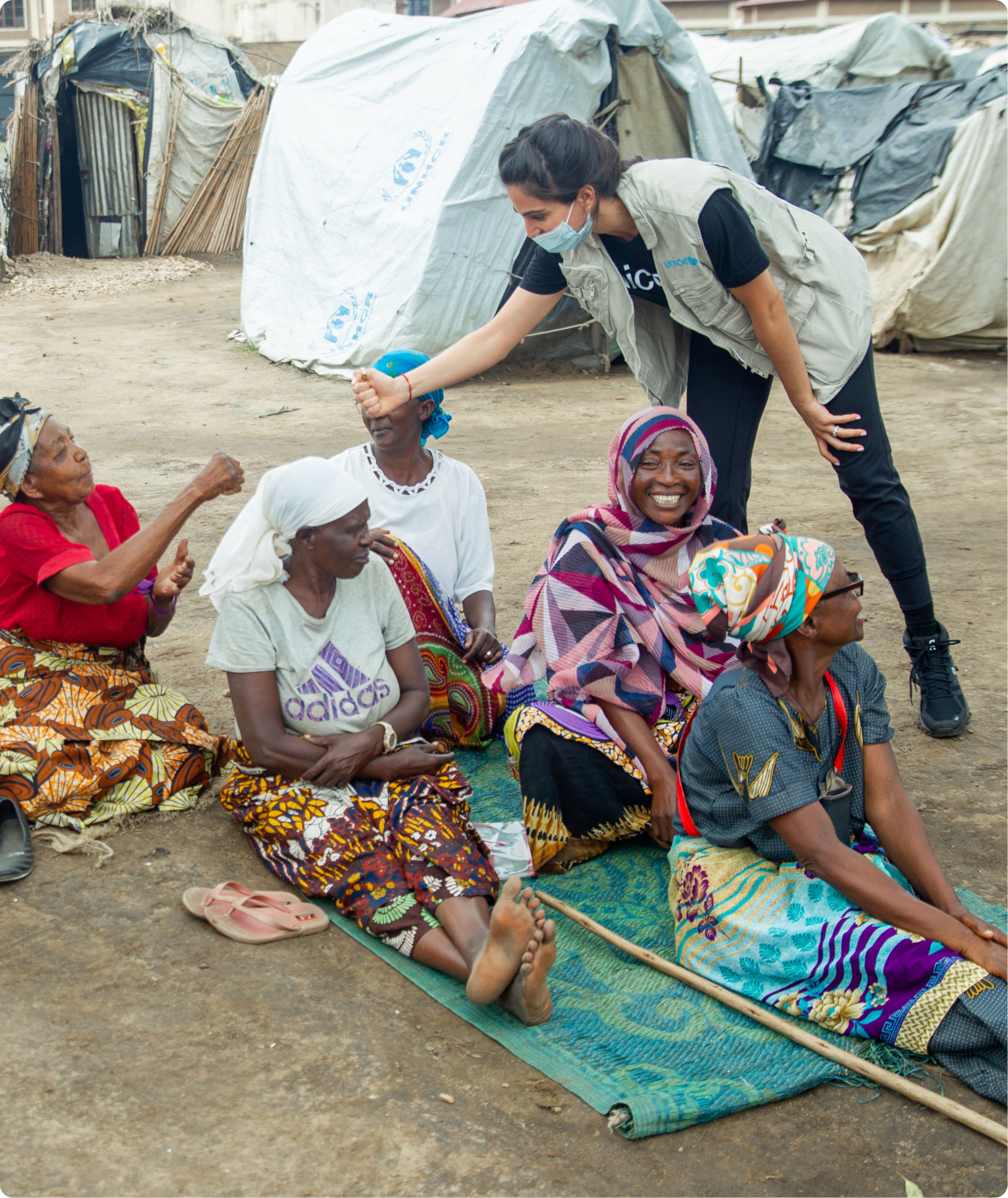 A woman with a blue mask giving a fist bump to an elderly woman sitting in a crowd of seated women, some wearing headscarves and colorful traditional clothing, on a makeshift mat outdoors with tents and makeshift shelters in the background.