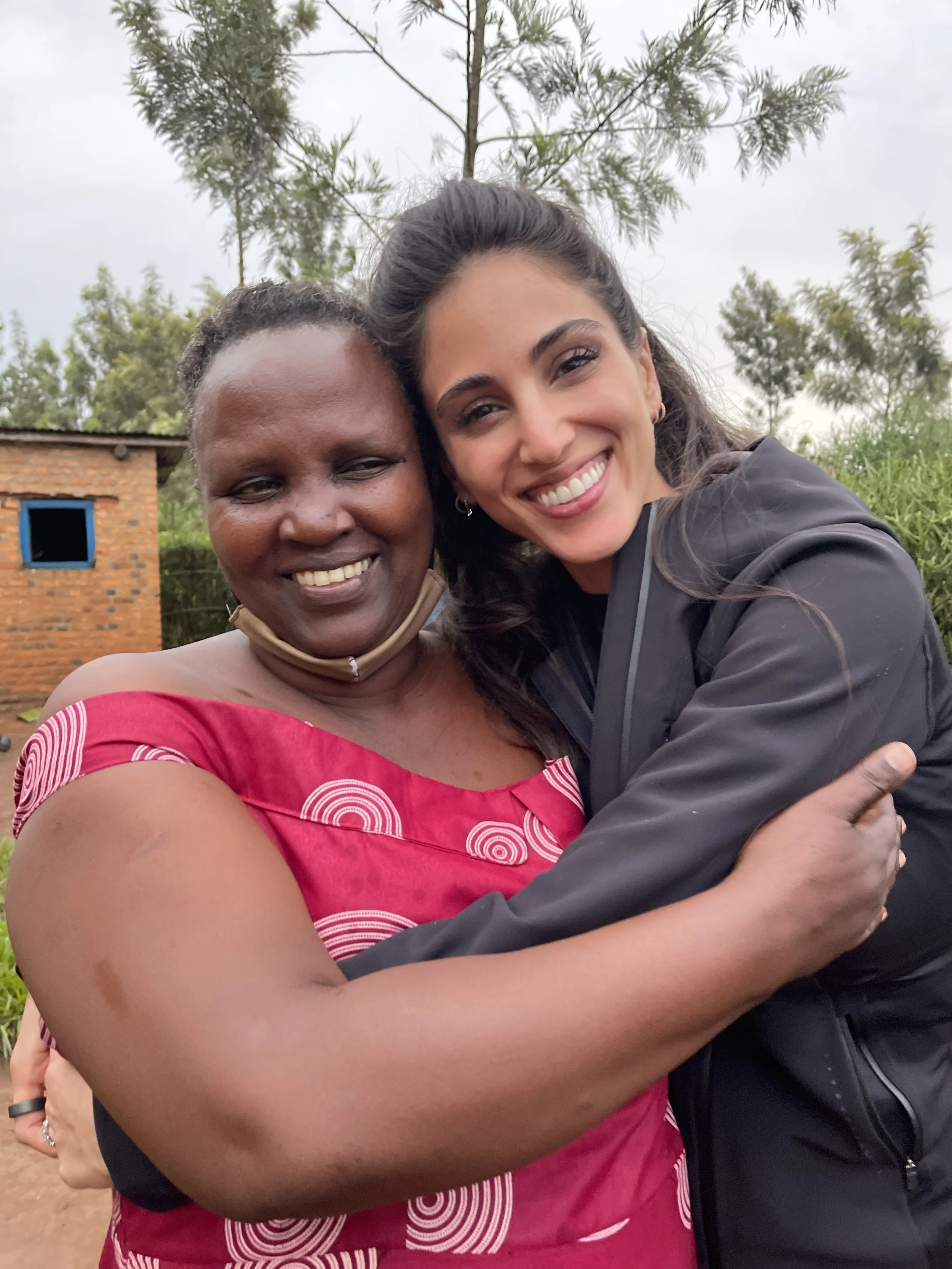 Two women hugging and smiling outdoors, with trees and a small brick building in the background.