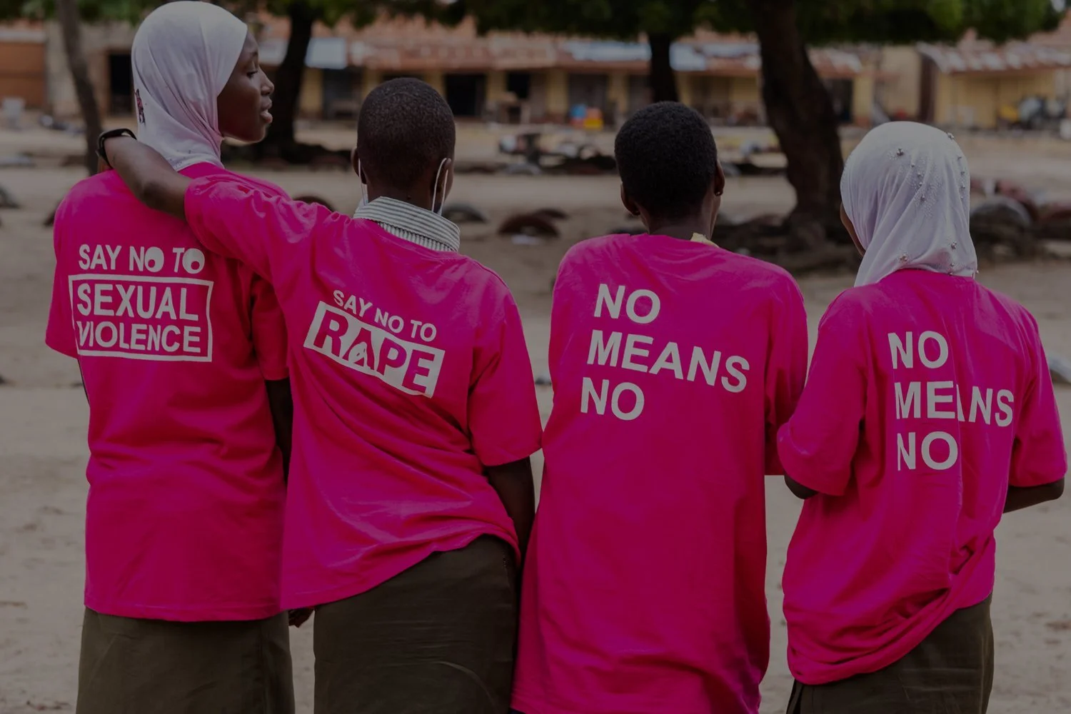 Group of children wearing pink shirts with anti-violence messages, standing outdoors near trees and buildings, seen from behind.