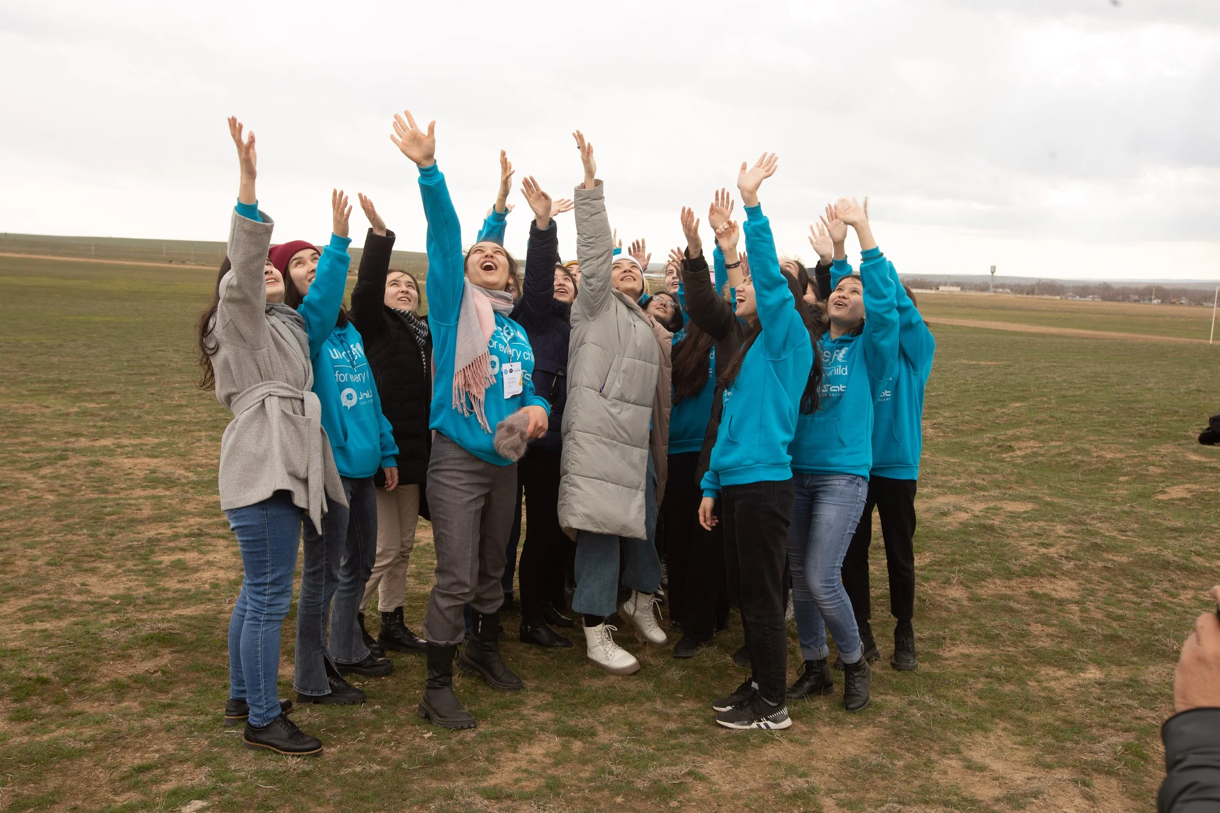 A group of young people standing outdoors on a grassy field, raising their hands up and smiling under a cloudy sky. Some are wearing blue jackets and are participating in an outdoor event or gathering.