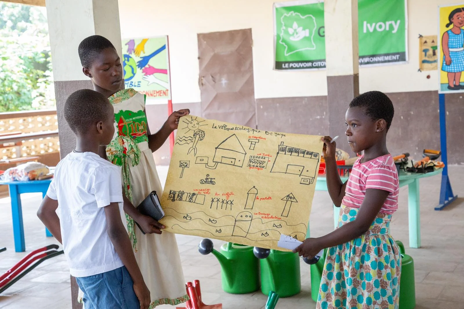 Three children are presenting a large illustrated poster about an ecological village to a teacher or a classmate. The poster includes drawings of houses, a playground, water, and animals, with French labels. The setting appears to be a classroom or educational space with colorful posters and toys around.