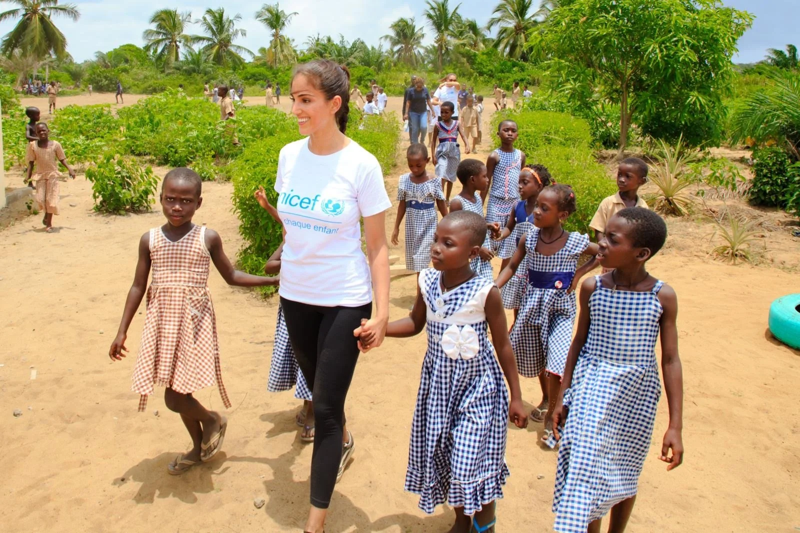 A woman wearing a UNICEF T-shirt walks hand-in-hand with young girls in school uniforms along a dirt path in a rural area with palm trees and greenery.