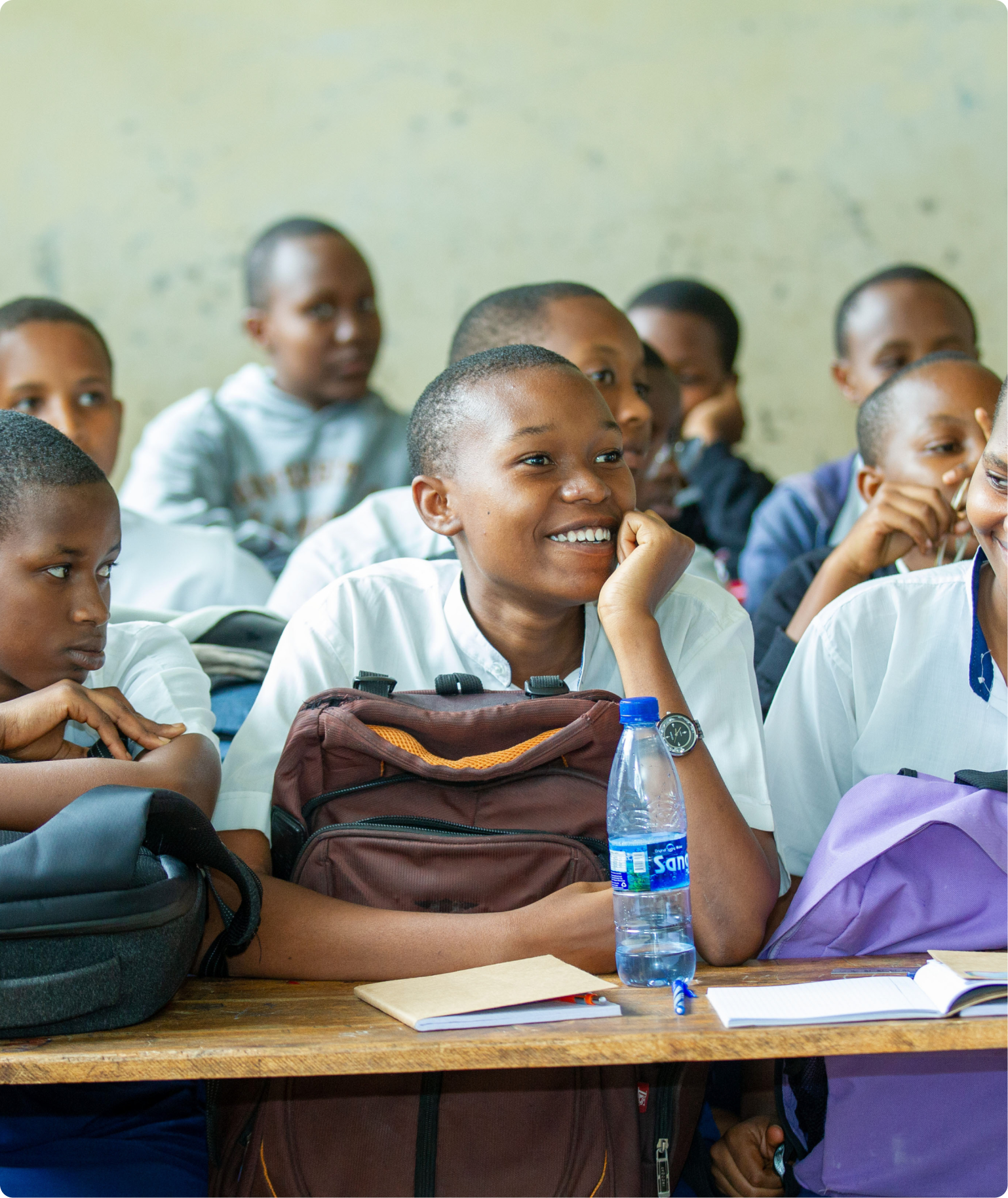 A group of school children sitting at desks in a classroom, some smiling and paying attention, with notebooks, backpacks, and water bottles on the desks.