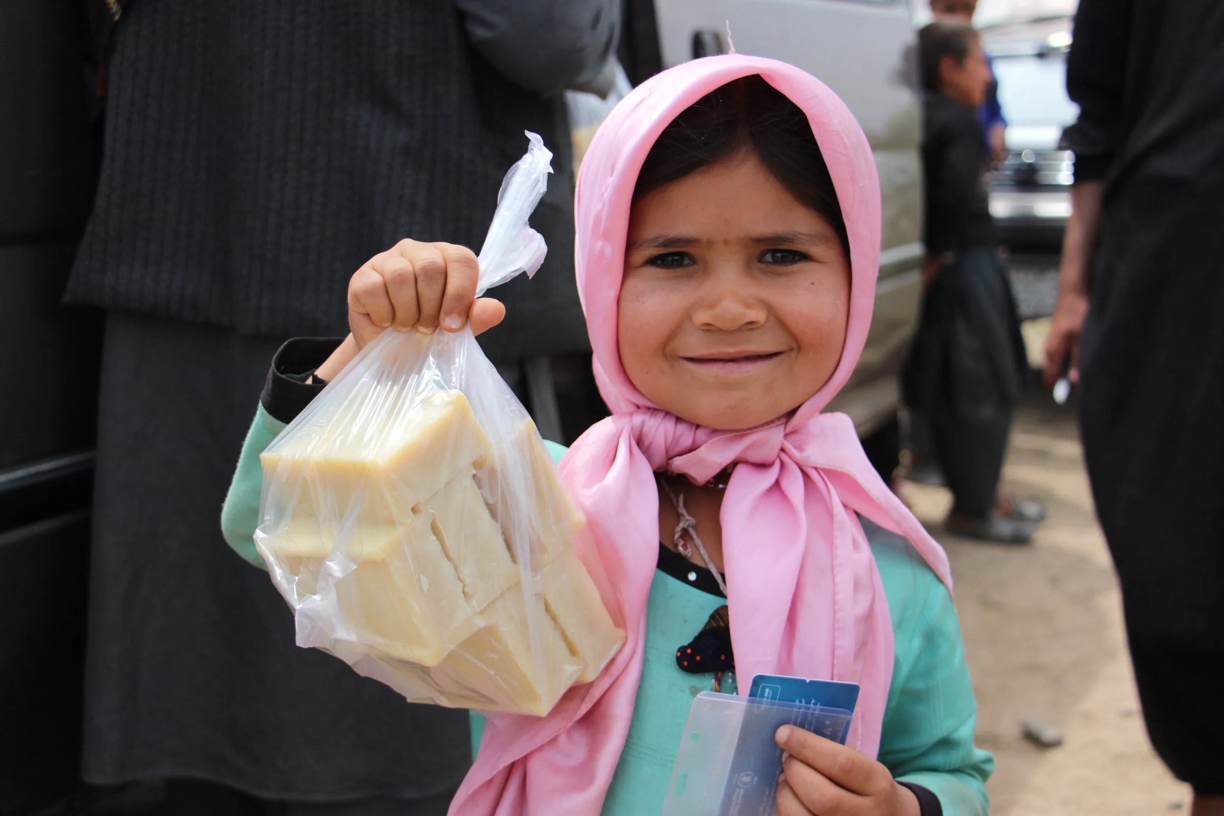 A young girl wearing a pink headscarf and teal jacket, holding a plastic bag with cheese and some cards, standing outdoors among people.